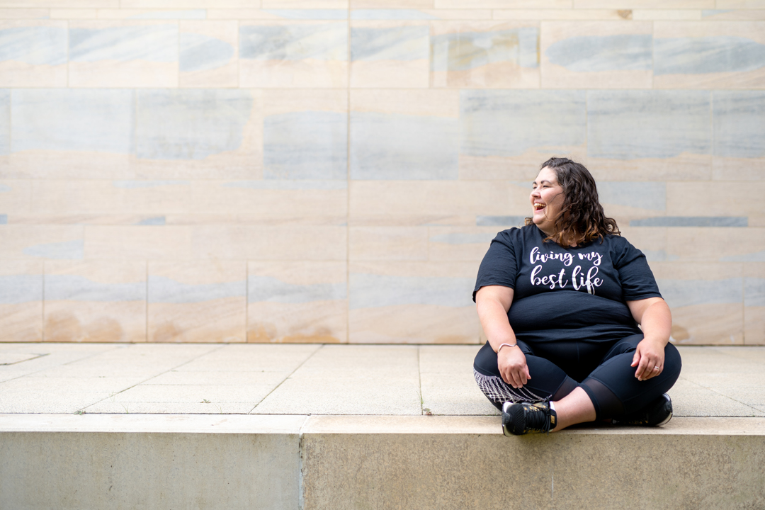 A plus-size woman with a T-shirt that says 'Loving my best life" takes a break by sitting on a marble ledge.