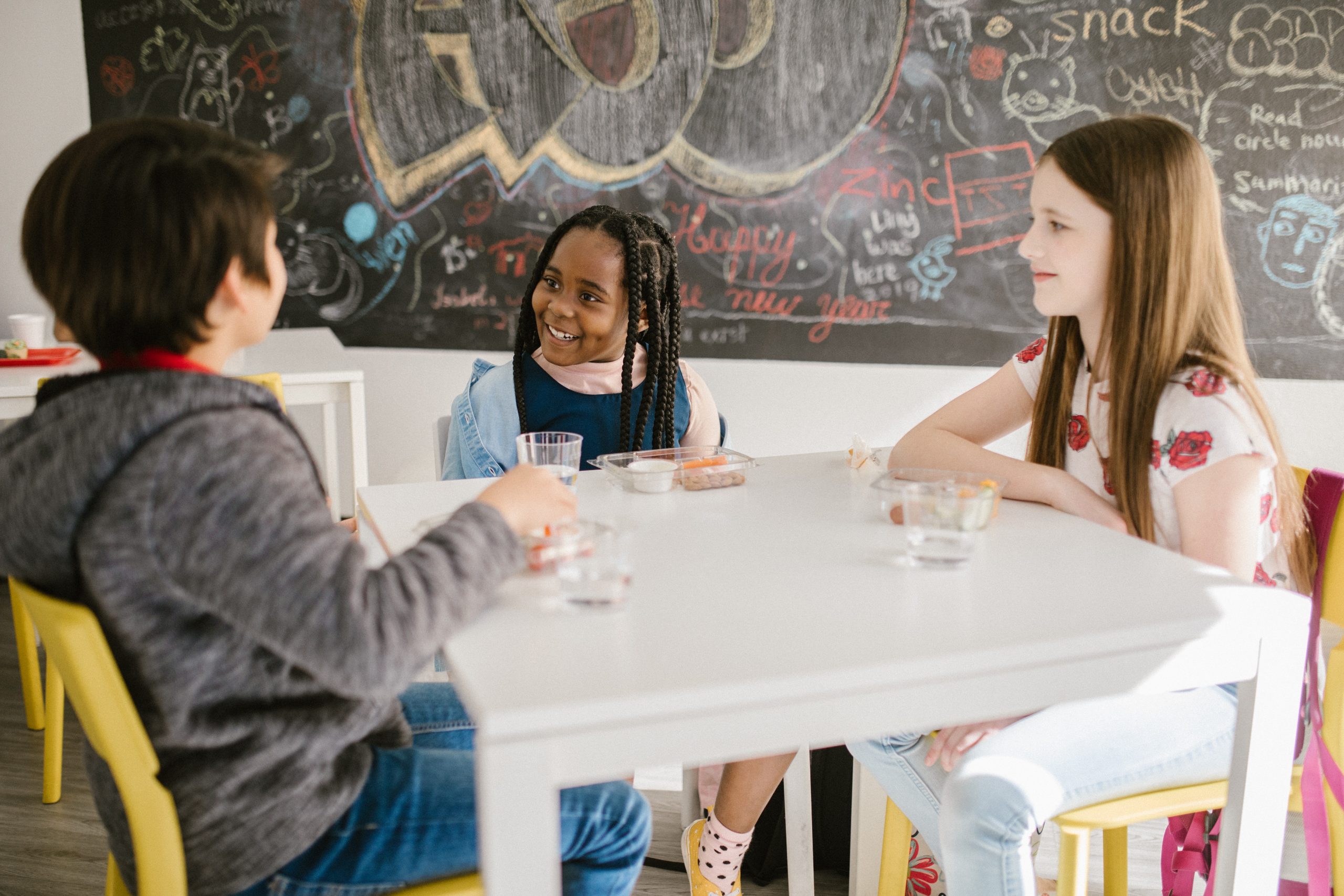 Three kids of different nationalities sitting around a white table in a classroom, happy and free to express themselves.