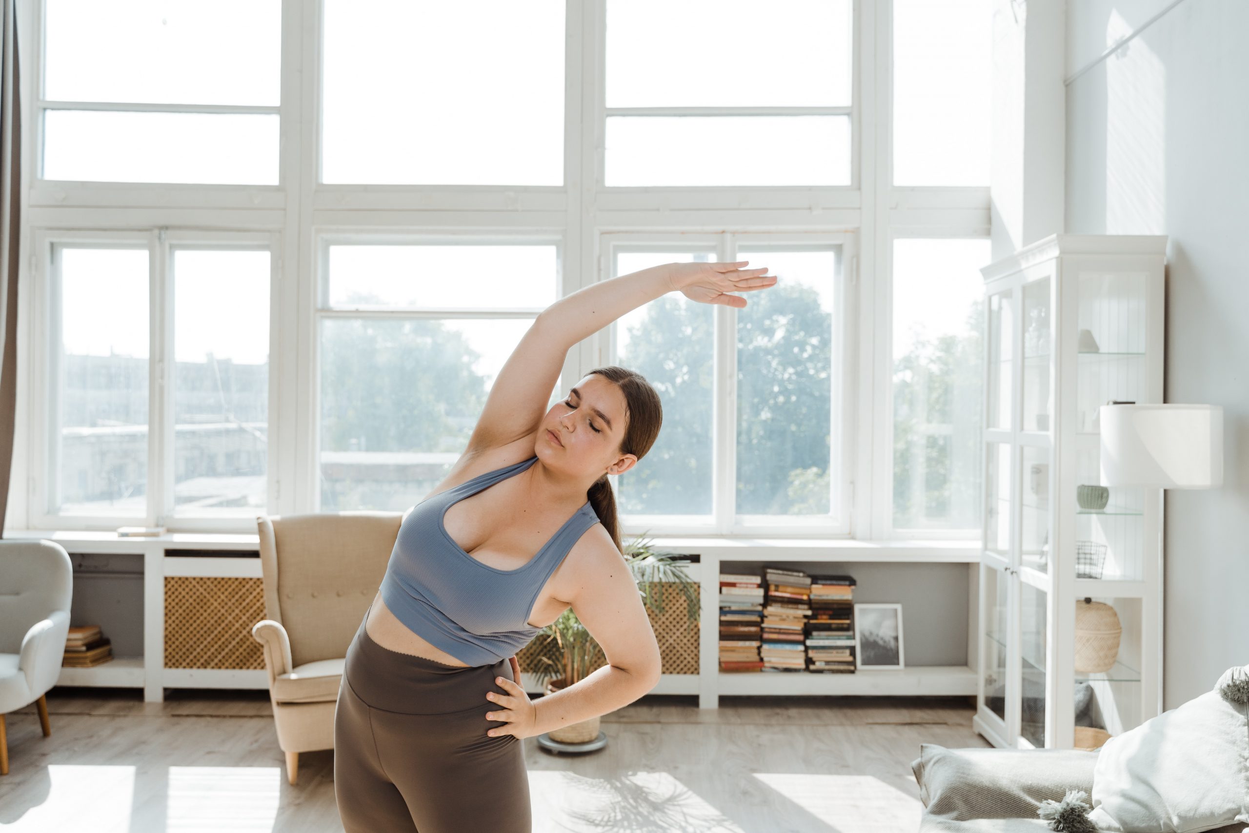 A young overweight woman practises yoga from home with light flooding from the windows behind her.