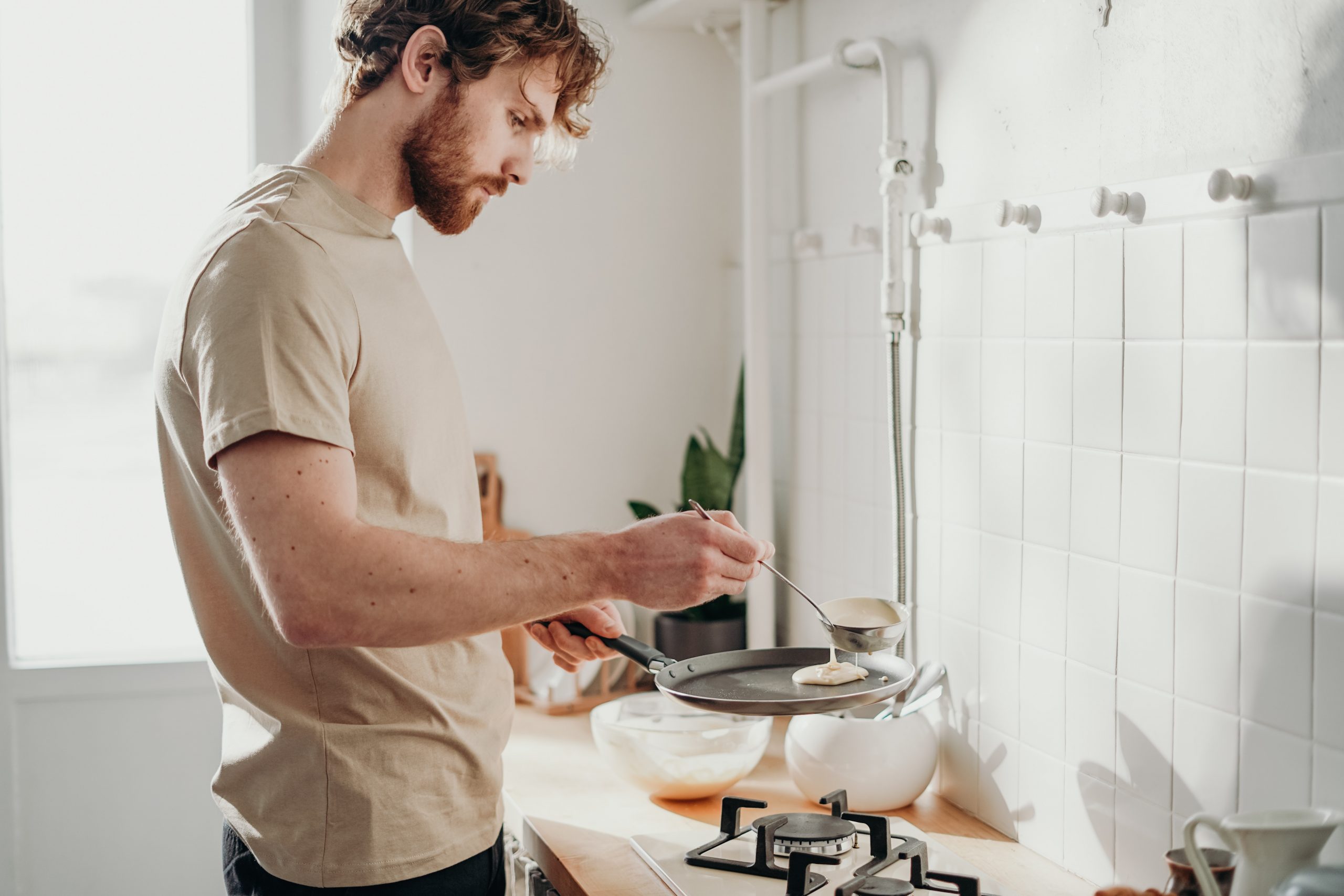 A young white man spoons a ladle of pancake mixture into a frying pan to make pancakes.