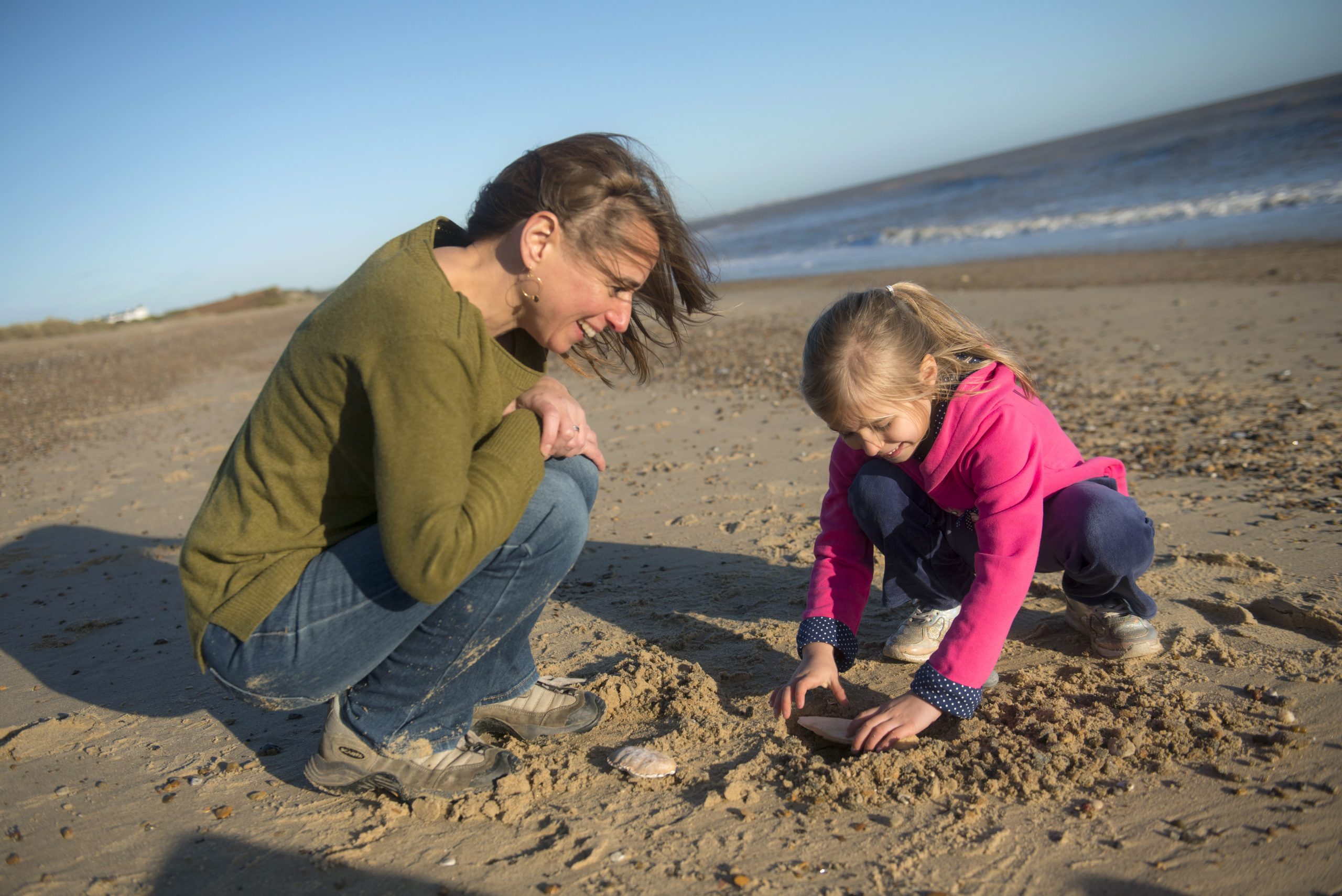 A mother crouches near her daughter playing in the sand on the beach. 