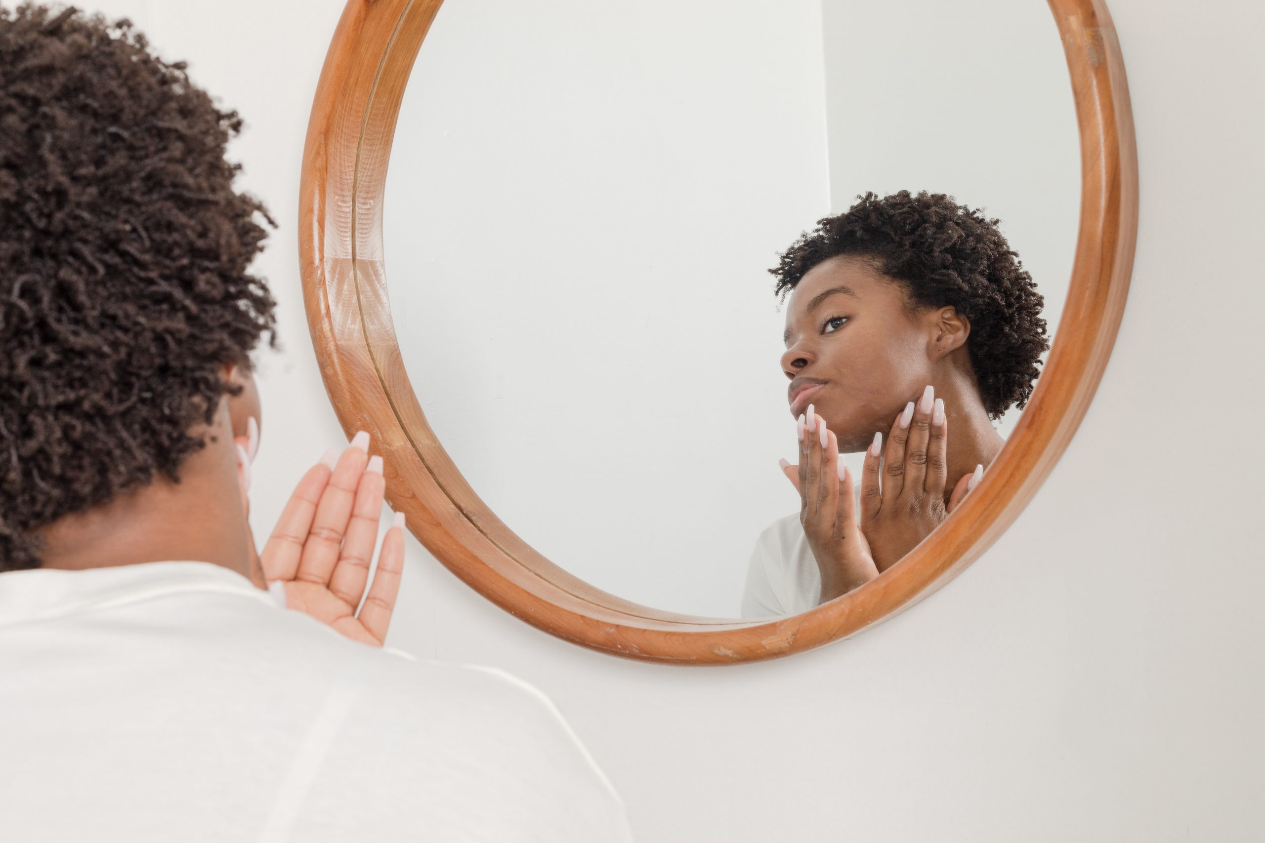 A young black woman applies her skincare routine in front of a wall mirror.