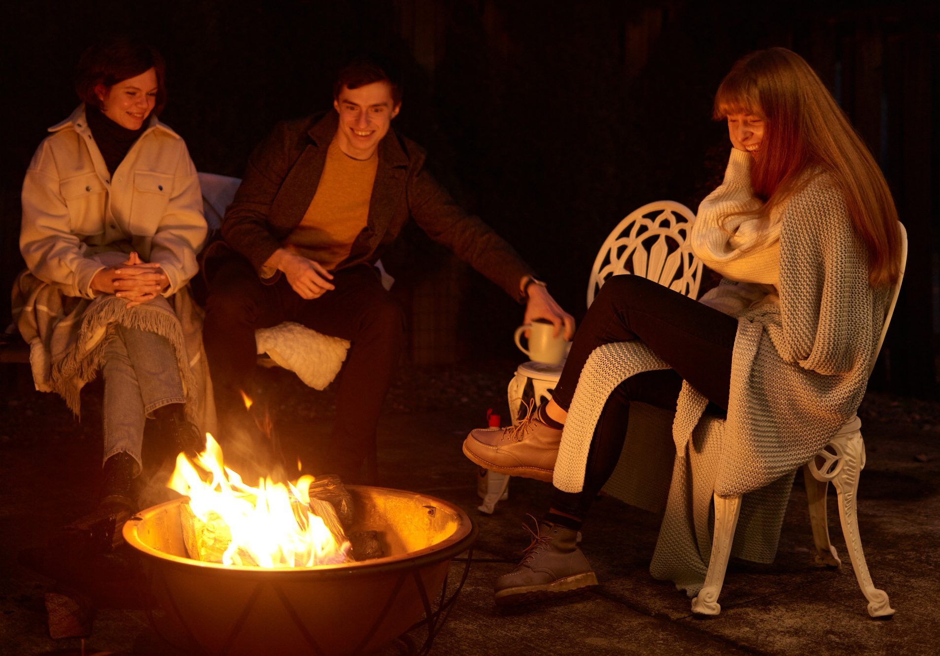 After dark, three young friends warm themselves around an outside firepit.