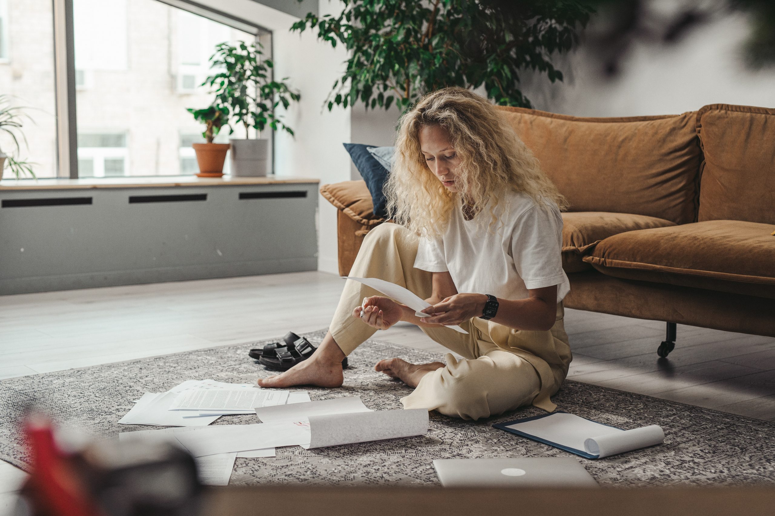 A blonde woman sorts through the paperwork of her finances, sitting on a rug on floor in her living room.