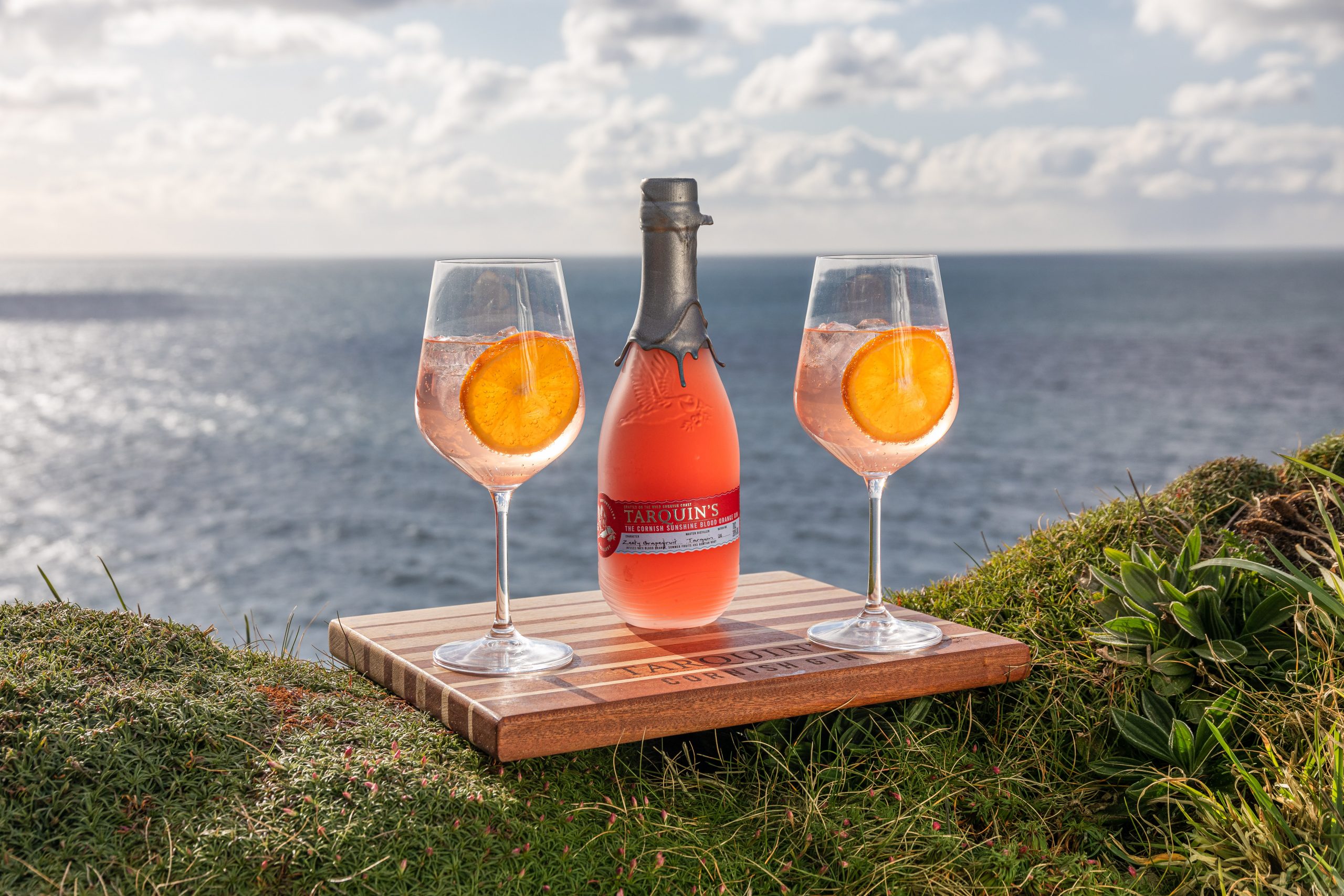 Two Blood orange sunshine spritz cocktails with a bottle of Tarquin’s Gin framed against the backdrop of the sea. 