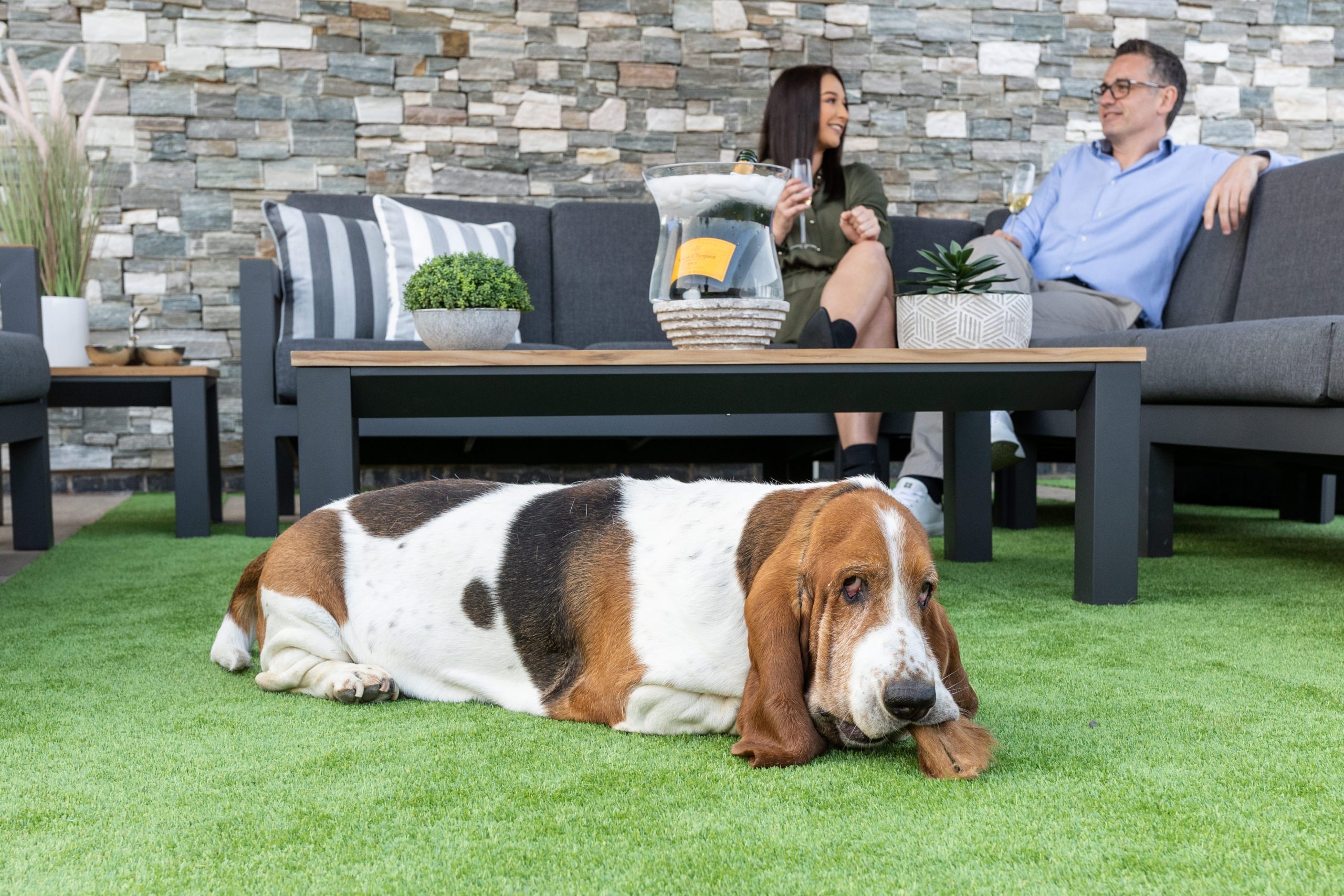 A couple relaxing on Sabai Living's 10-seater garden furniture set, with their pet Basset Hound in front of them.