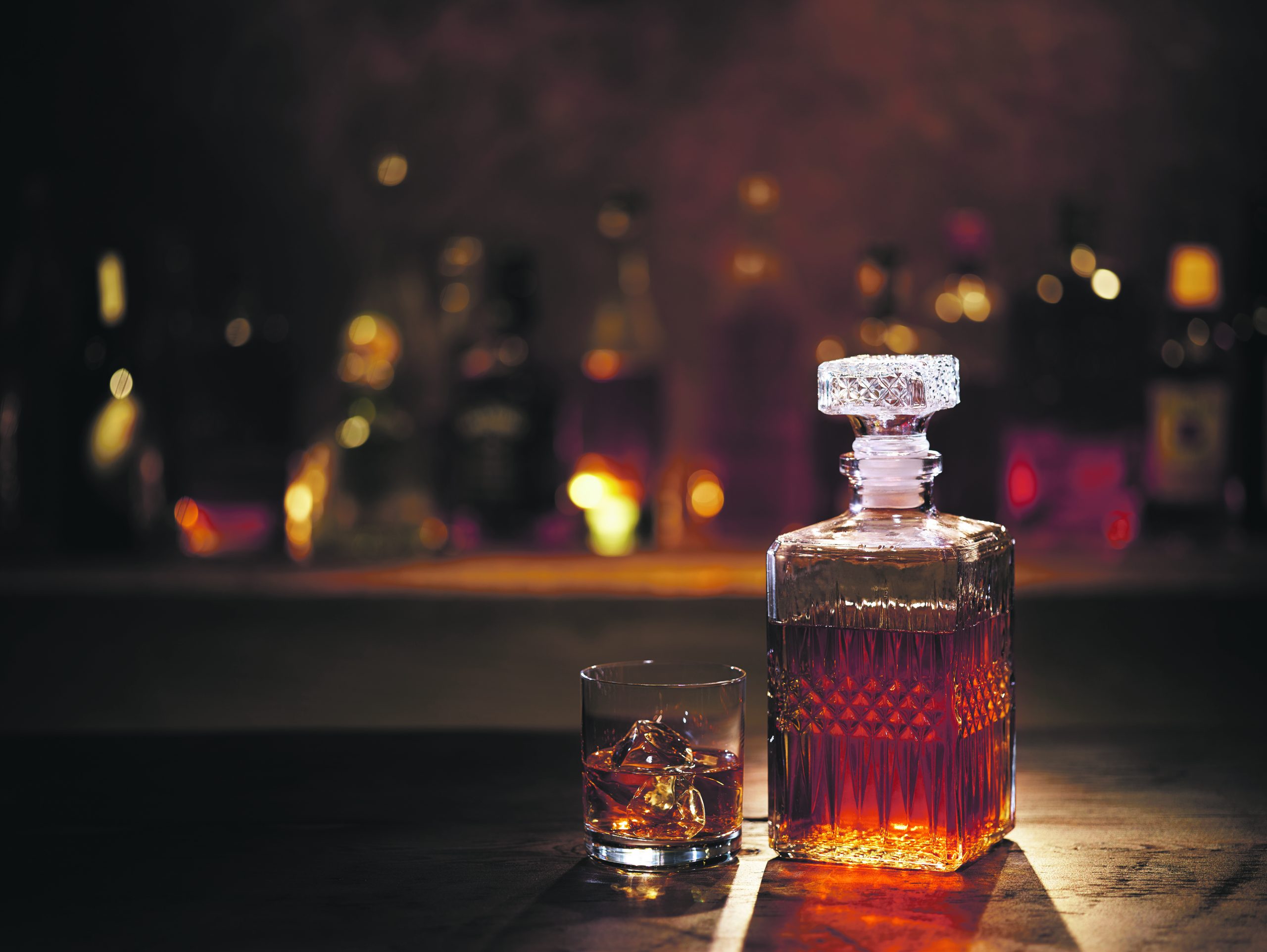 Backlit, a glass of whisky and decanter on the counter of a bar.