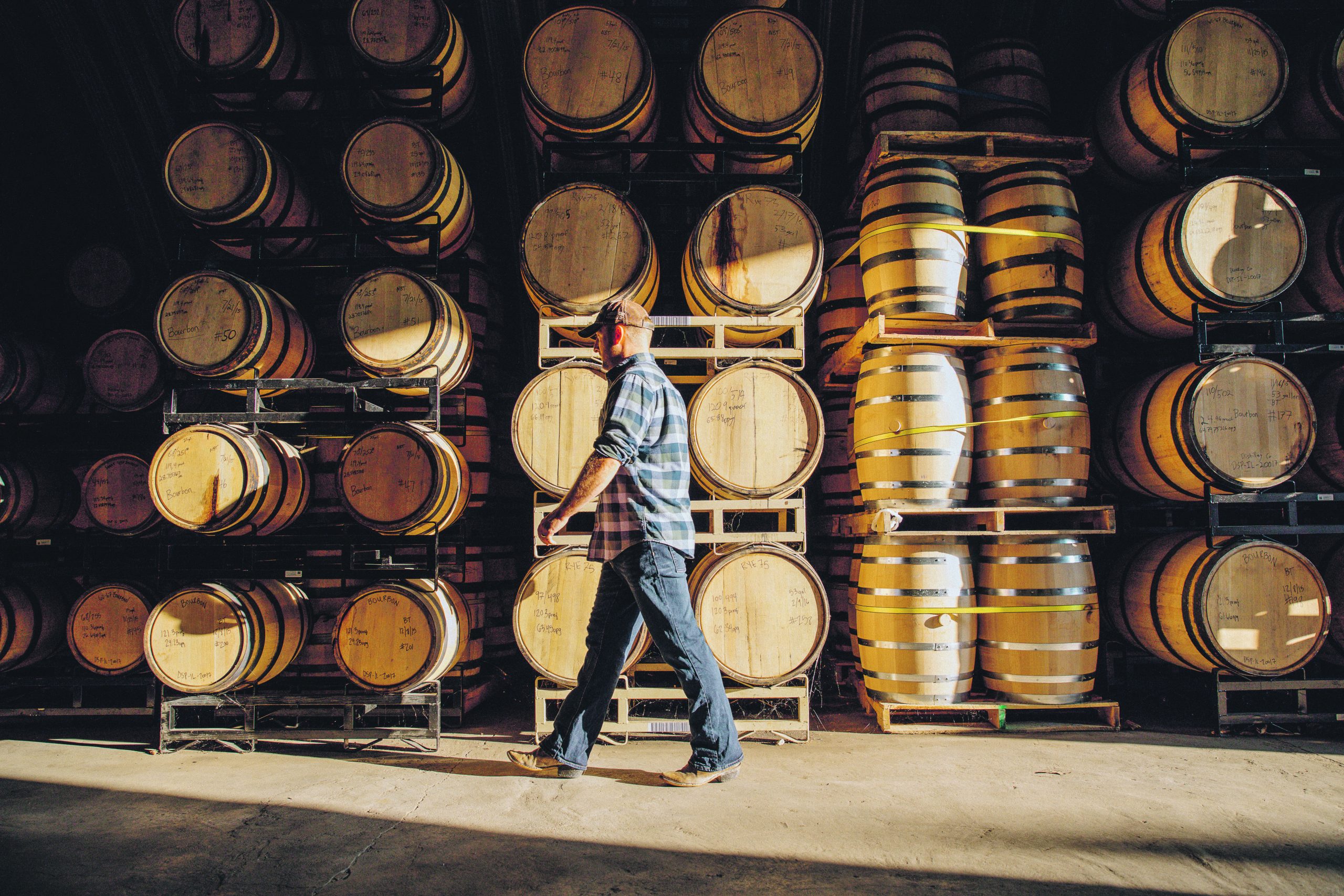 A worker walking past a wall of kegs inside a whisky distillery.