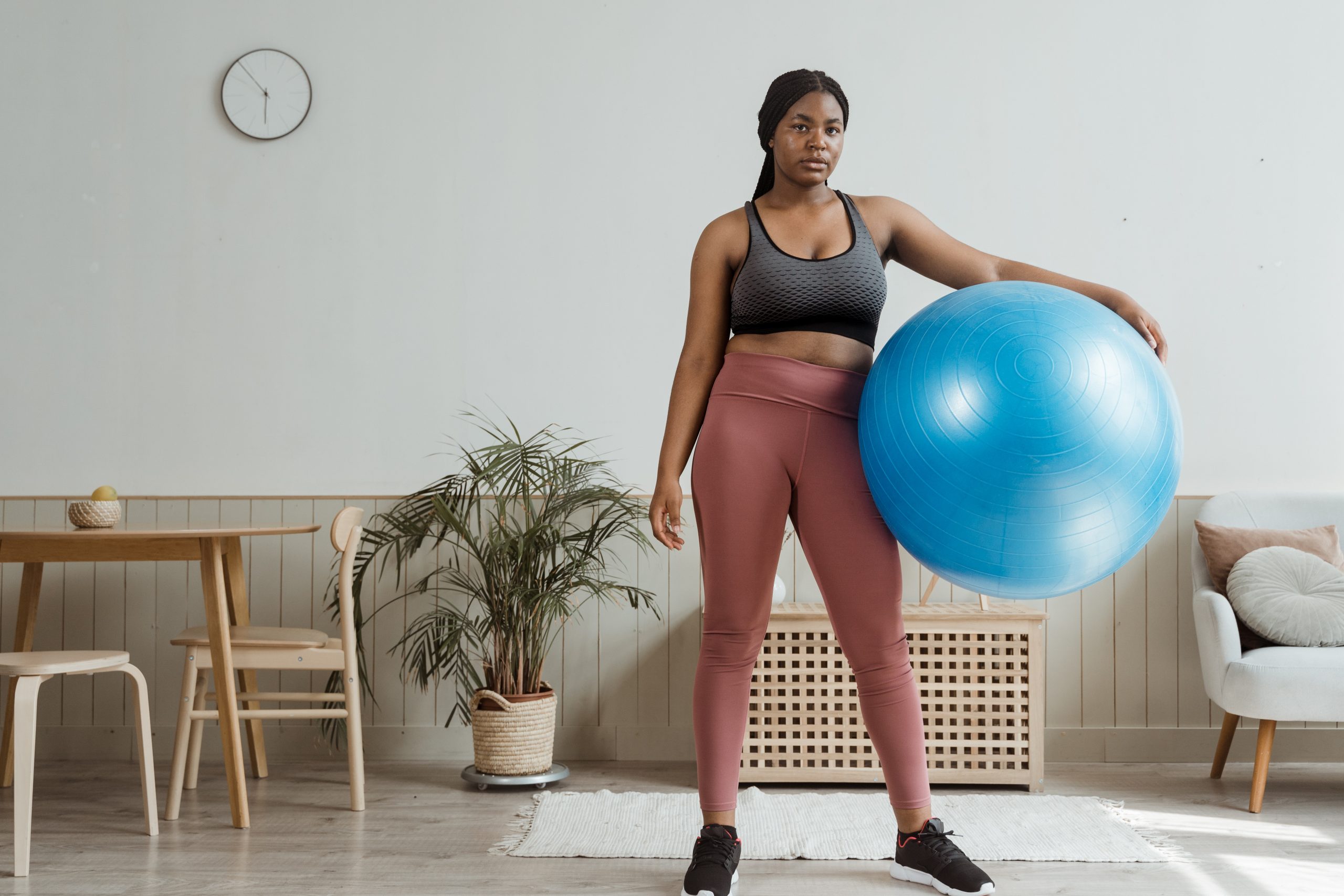 Holding a blue gym ball, a young black woman prepares to train in her living room.