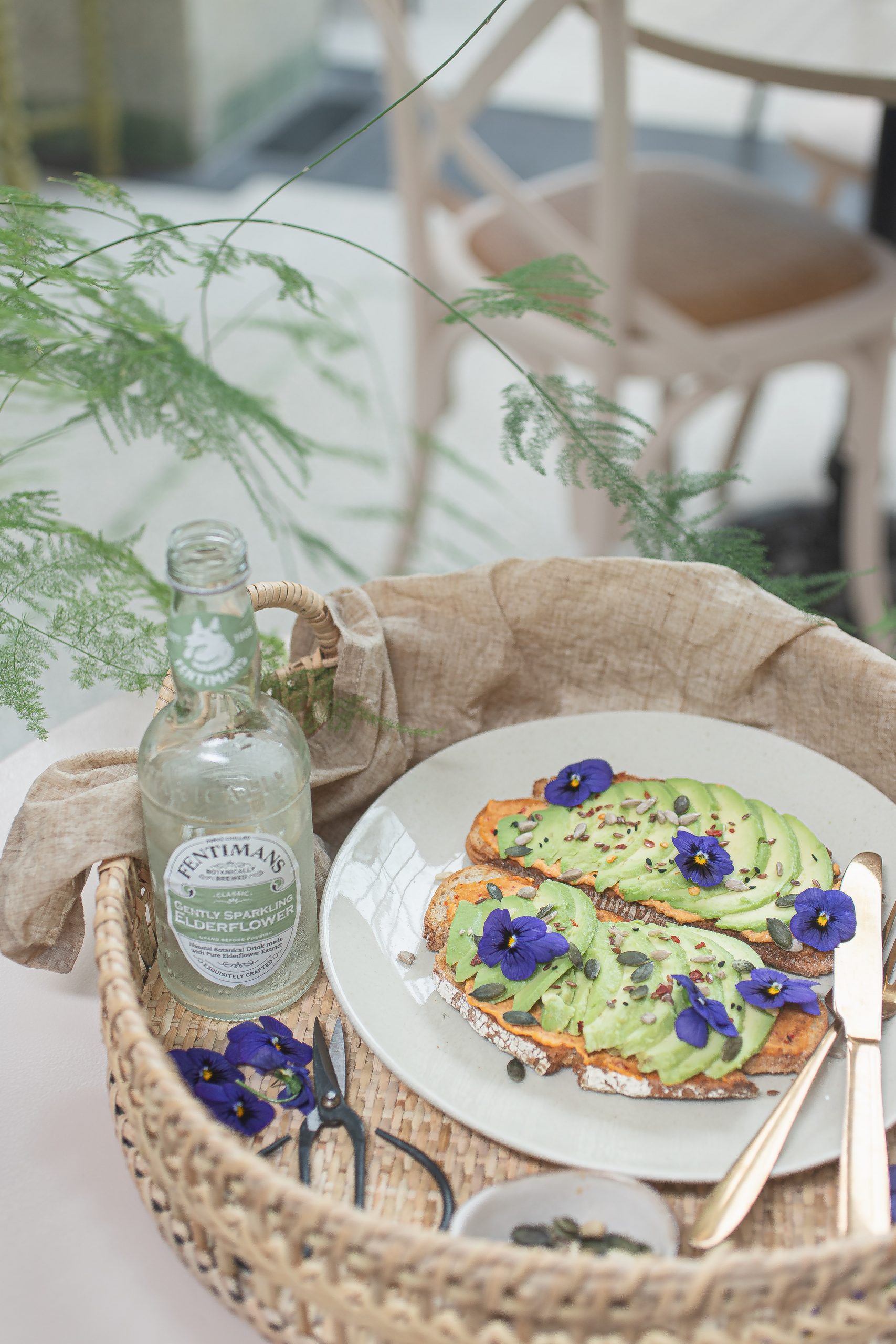 Sliced rye bread with avocado and violets, next to a bottle of Elderflower drink.