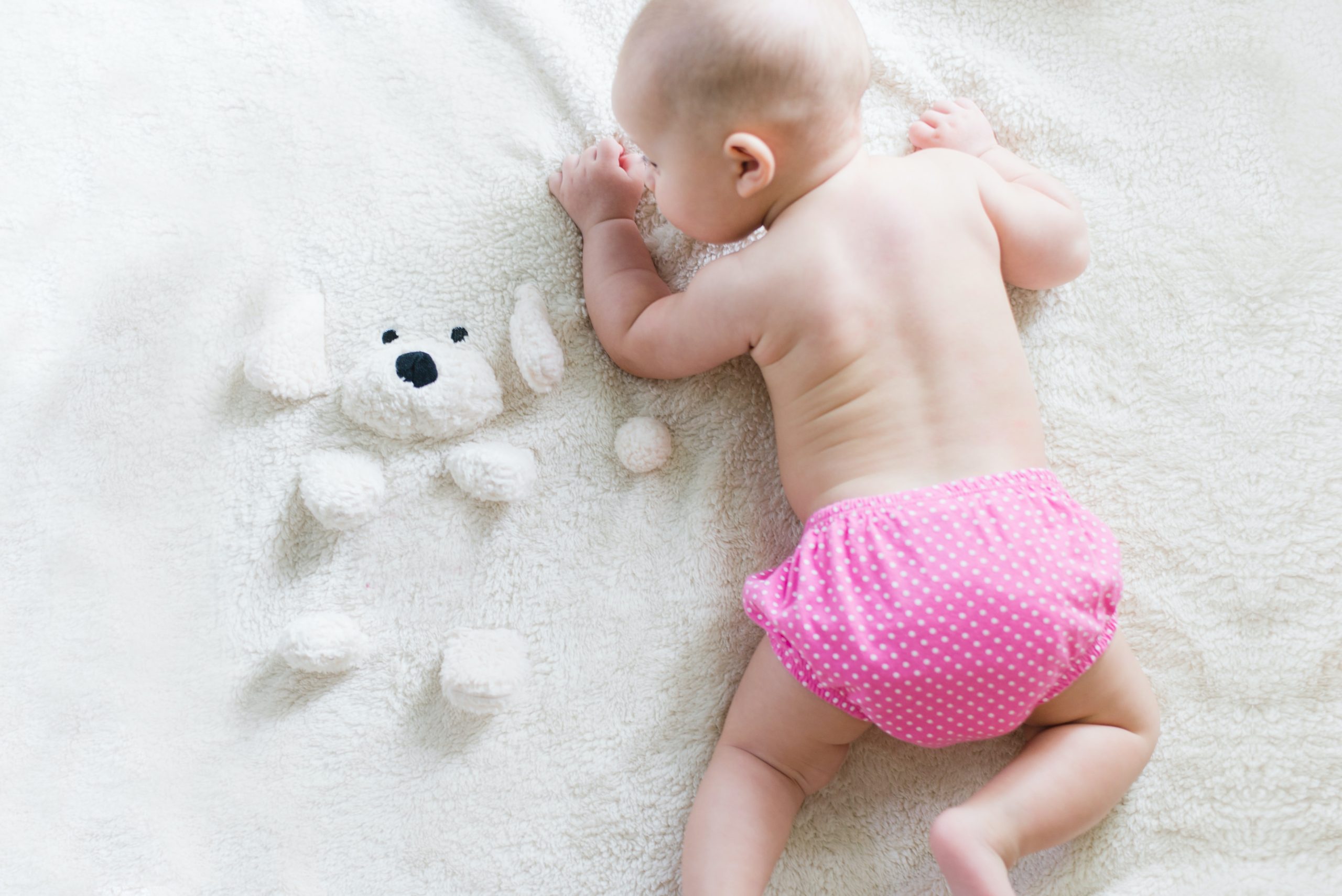 A baby crawling in a pink reusable nappy next to a teddy bear emerging out of a towel.