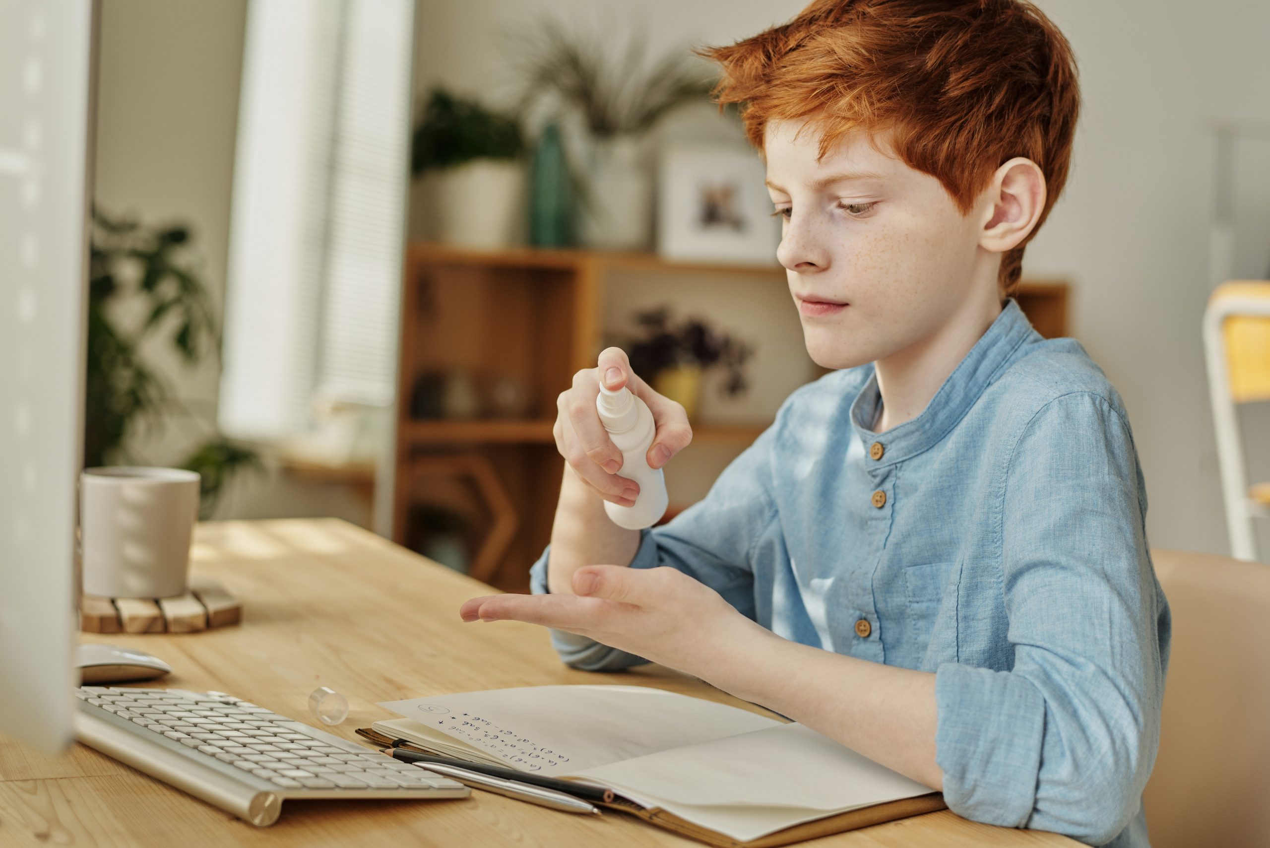 A young boy sanitising his hands with a spray before working on his homework on the computer. 
