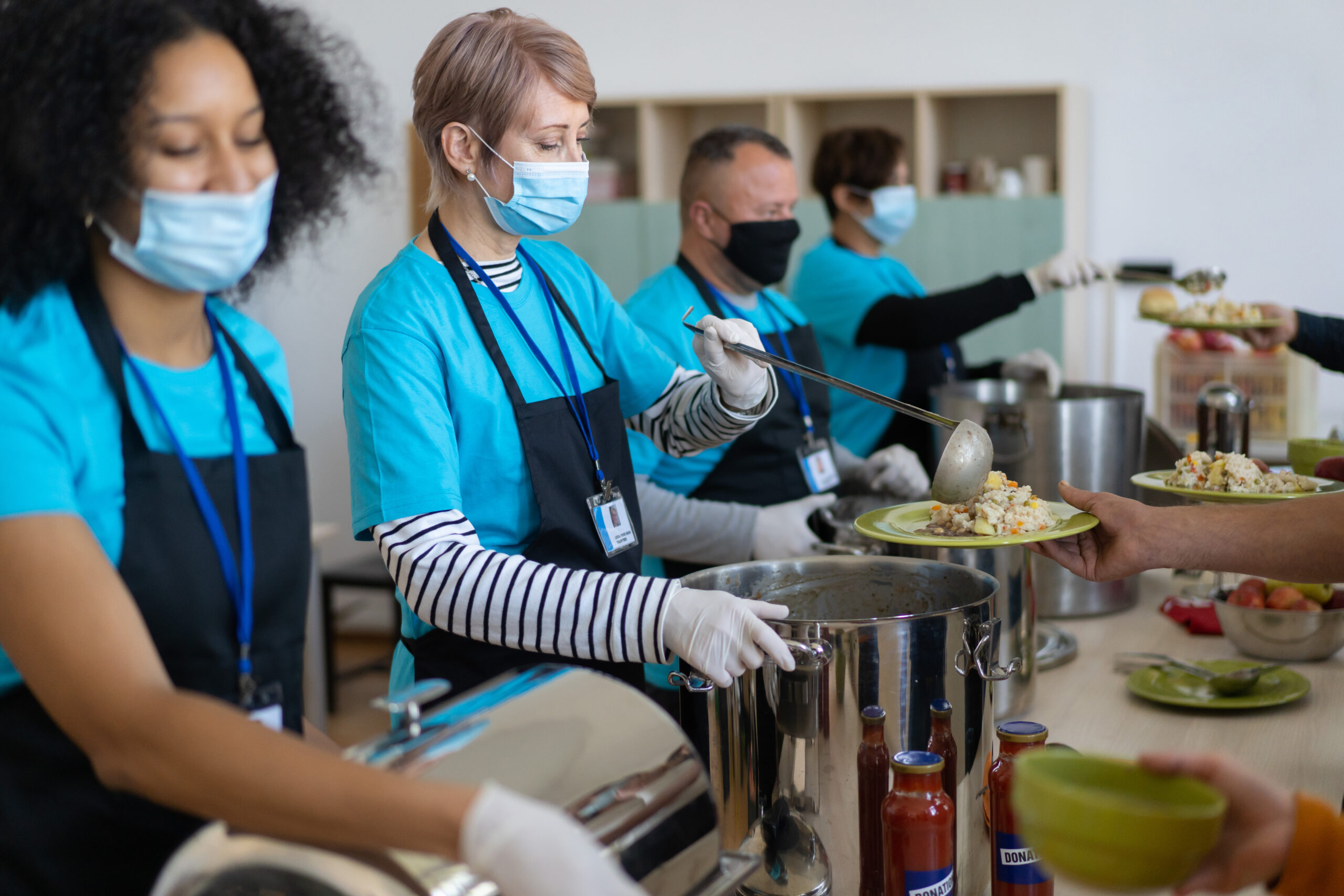 A group of volunteers serving donated food, lading food onto plates to disadvantaged people, wearing protective face masks, aprons and protective gloves 