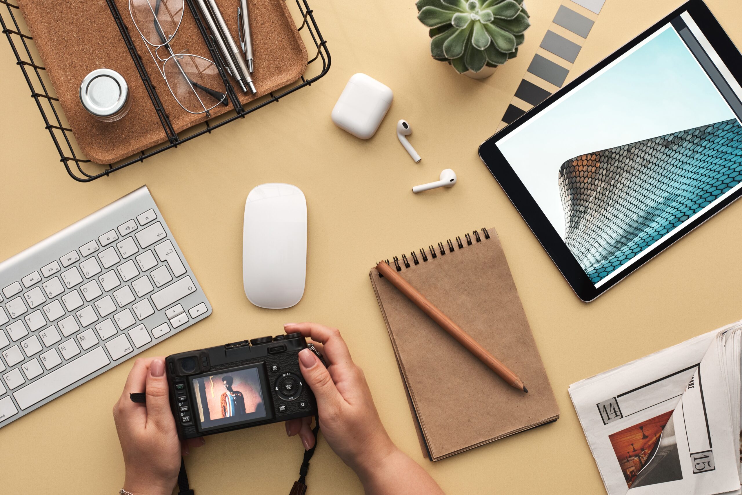 Detail of a man's hands scrolling through the pictures on his camera at a desk with other tech gadgets and a notebook.