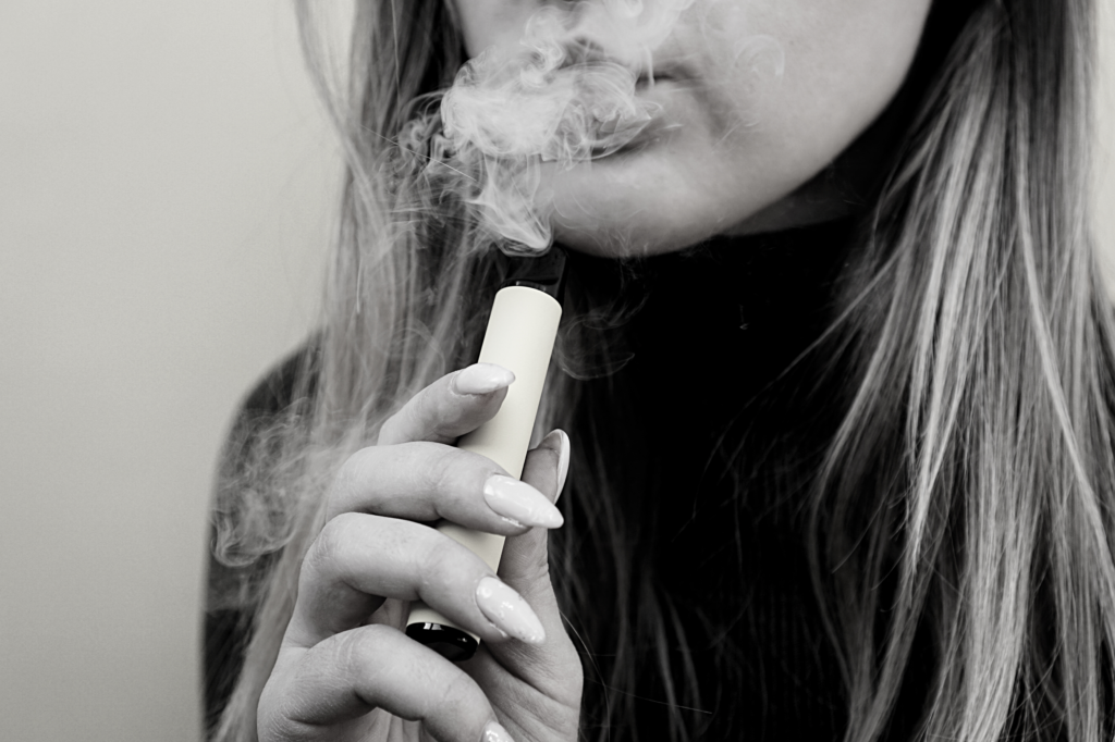 A black-and-white closeup of a young woman vaping an Elf Bar as she breathes out a cloud of smoke, the picture framed to hide her identity.