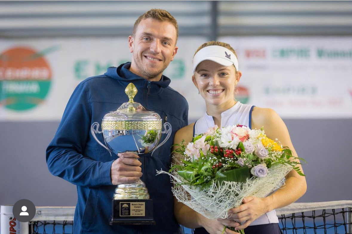 Tennis coach Alex Ward and Katie Swann smile for the camera, holding a trophy and a bunch of flowers.