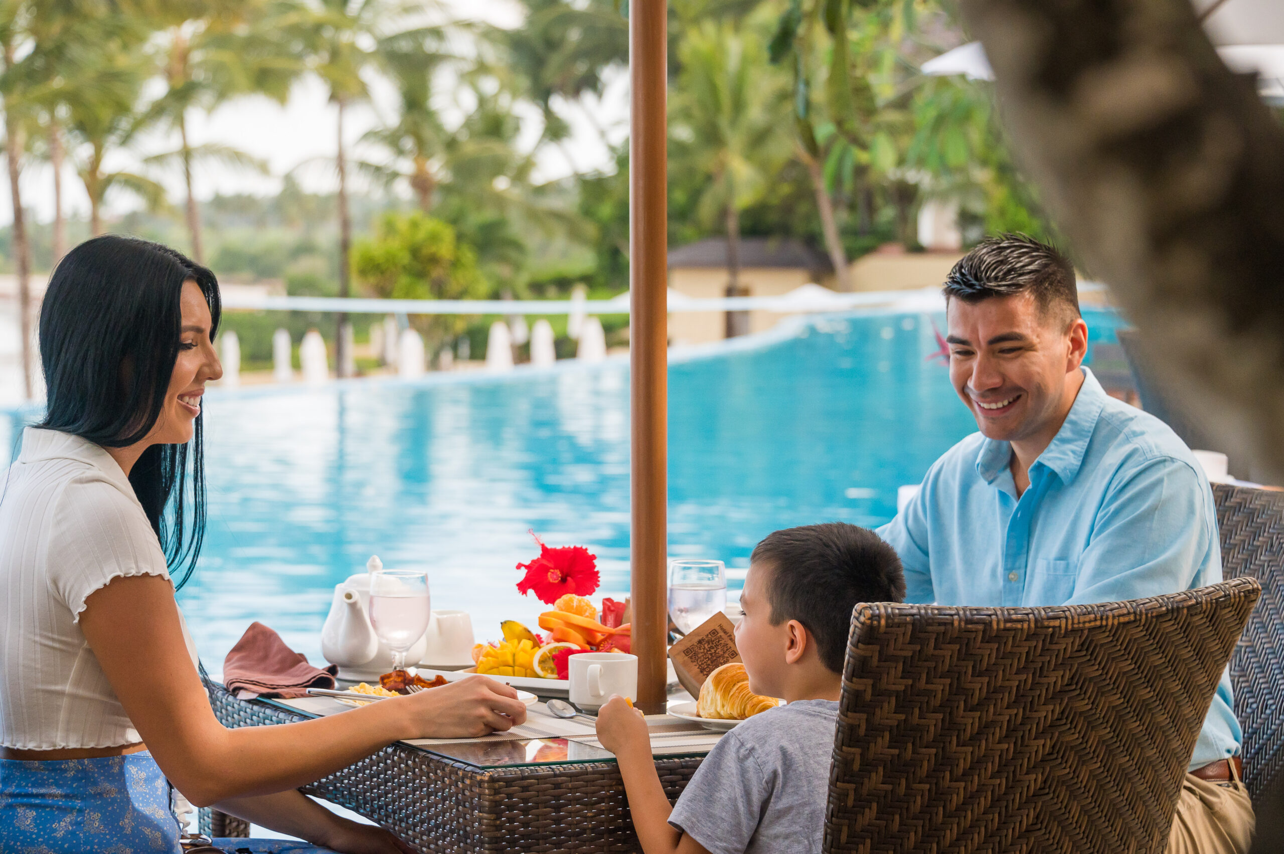 A couple sit with their son eating breakfast outside by a pool