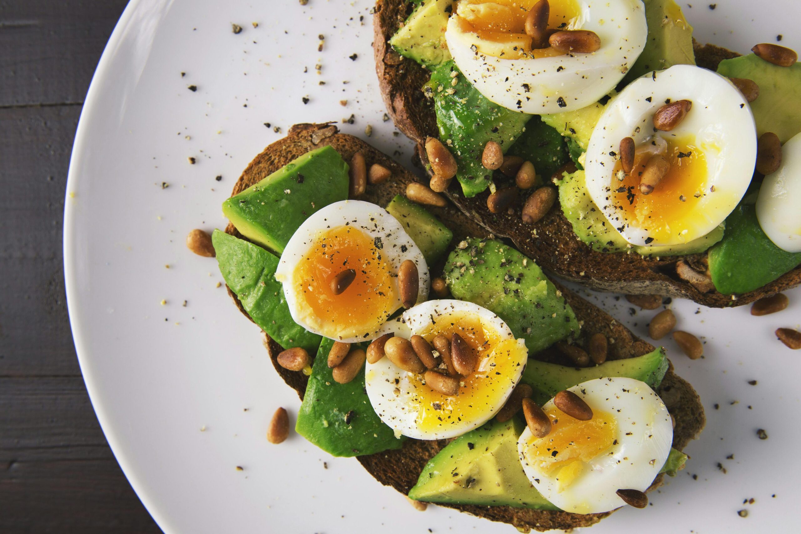 Avocado on toast with halved boiled eggs and nuts sprinkled, on a white plate