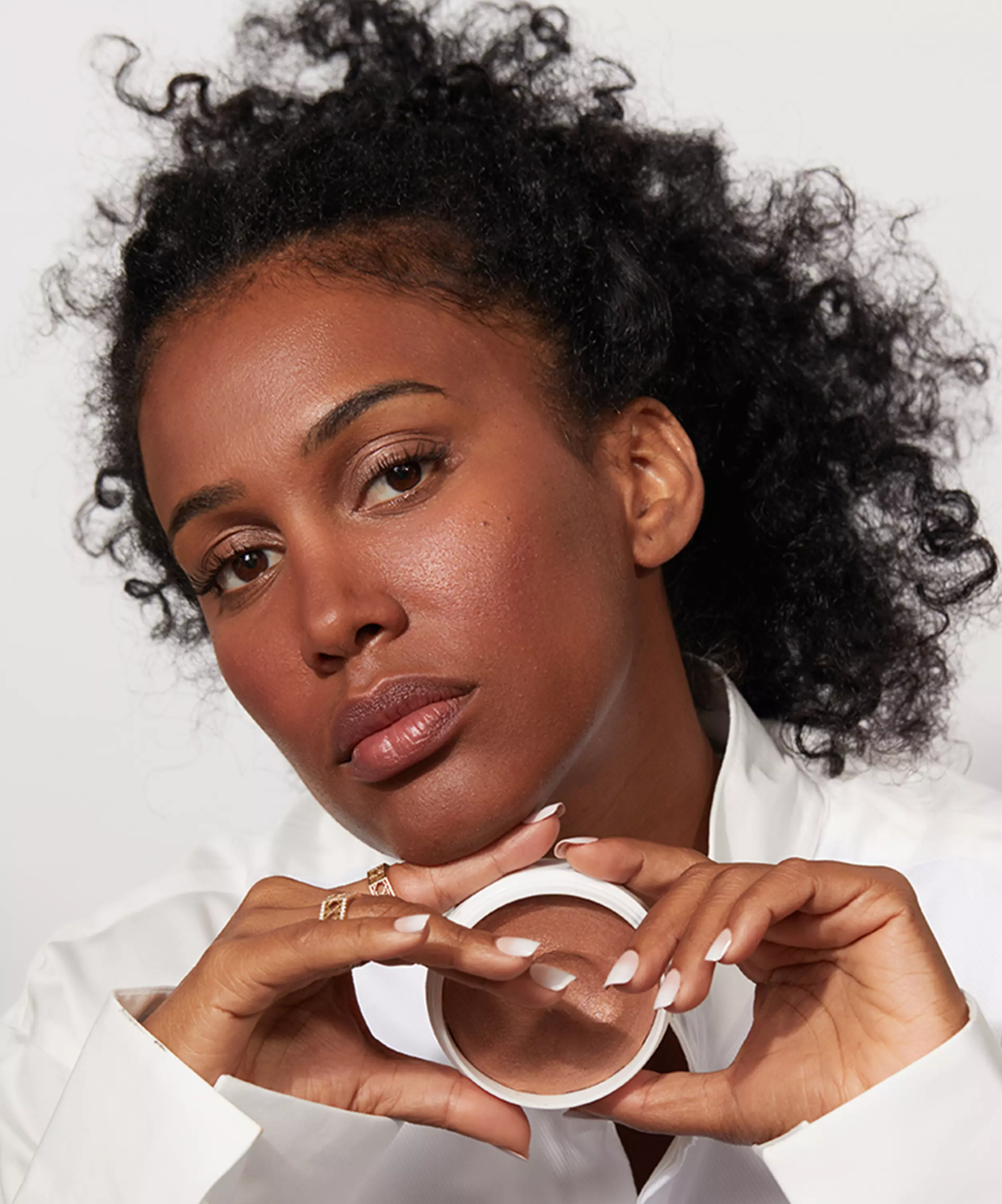 Close up of a black woman holding an open pot of Jones Road Miracle Balm in shade Magic Hour which is on her skin