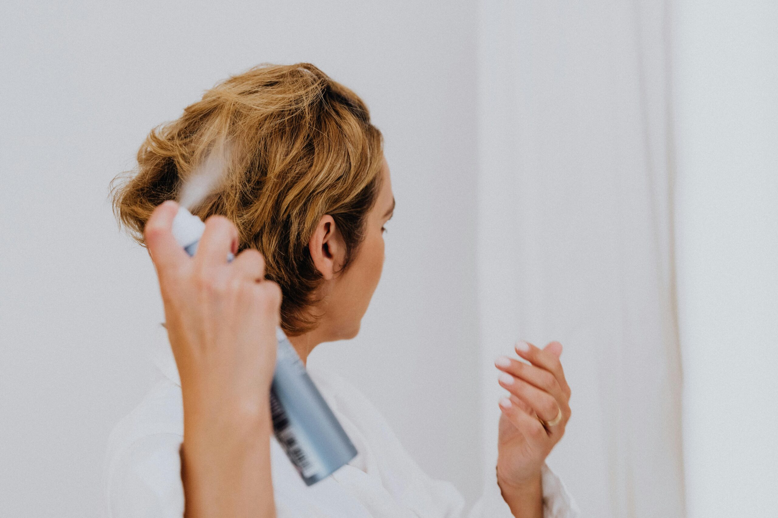 woman sprays dry shampoo onto the back of her short choppy hair