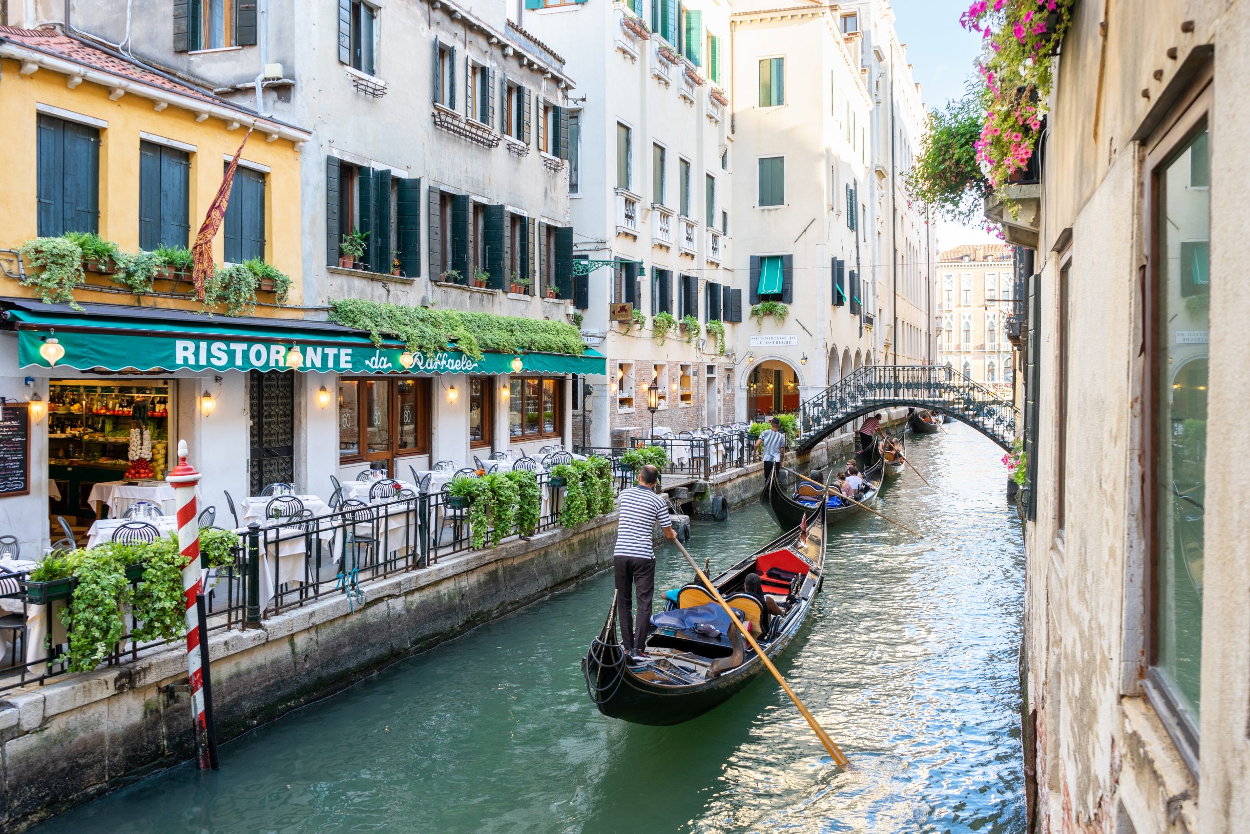 Two gondolas sail down a Venice canal, passing homes and restaurants on the bank