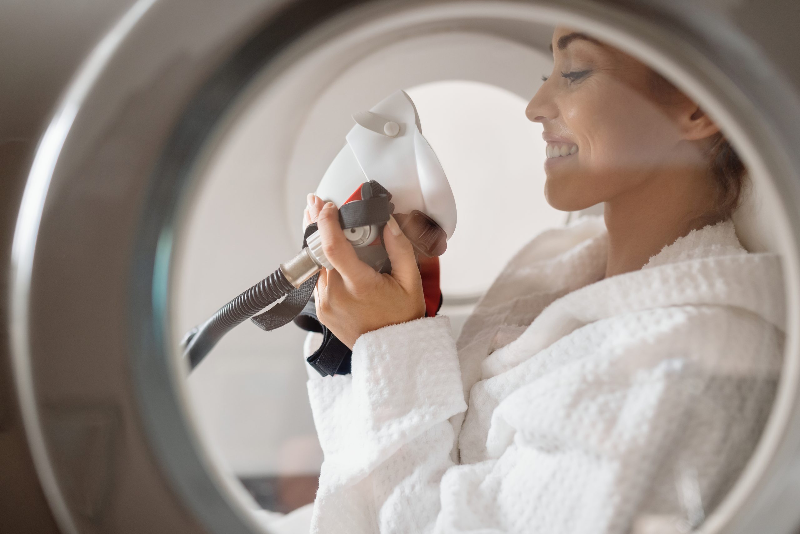 A smiling woman brings an oxygen mask to her face during her hyperbaric oxygen therapy treatment at a health spa.