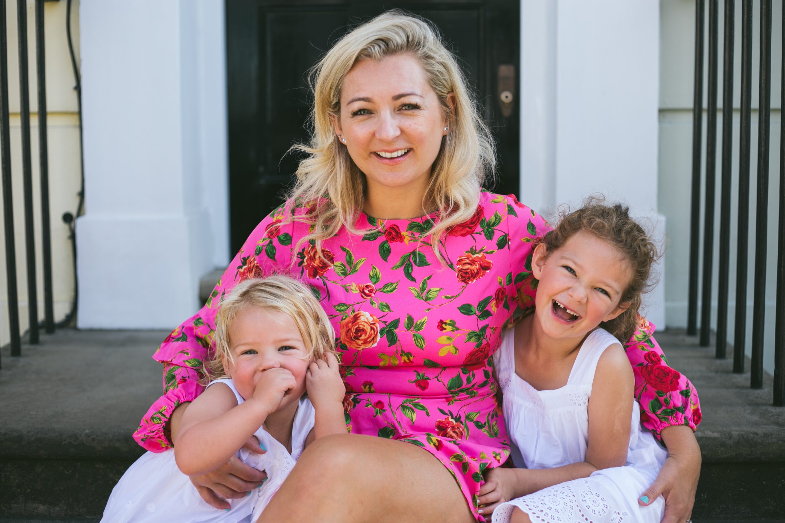 Rosey Davidson, founder of Just Chill Baby Sleep, poses in a pink dress with two of her daughters on her front porch
