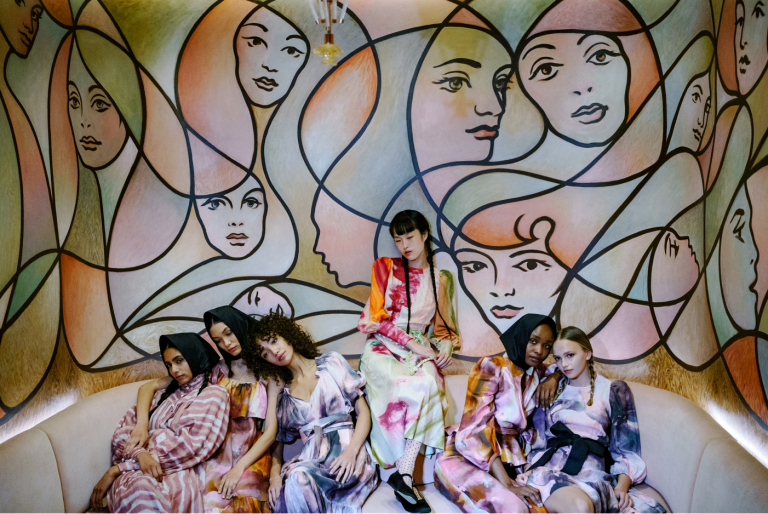 Seven international young women huddled against a mural of women's hairstyles.