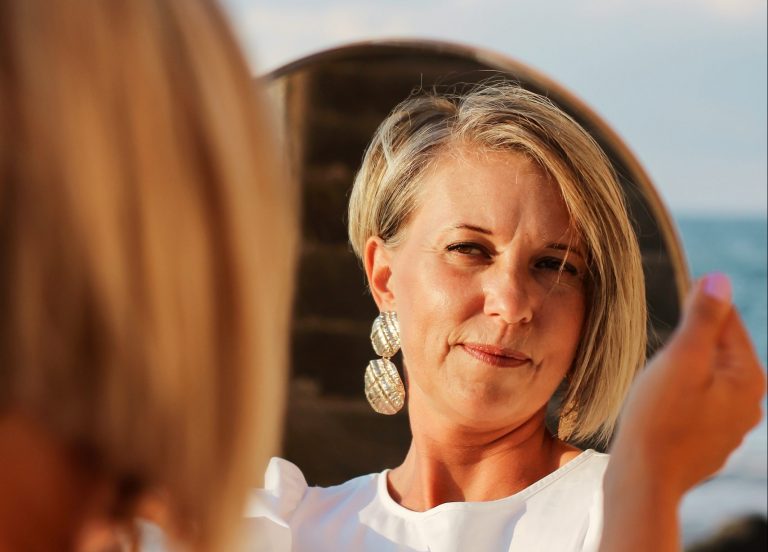 A woman in her fifties looks at herself in a mirror, mindful of the changes in her skin during menopause.