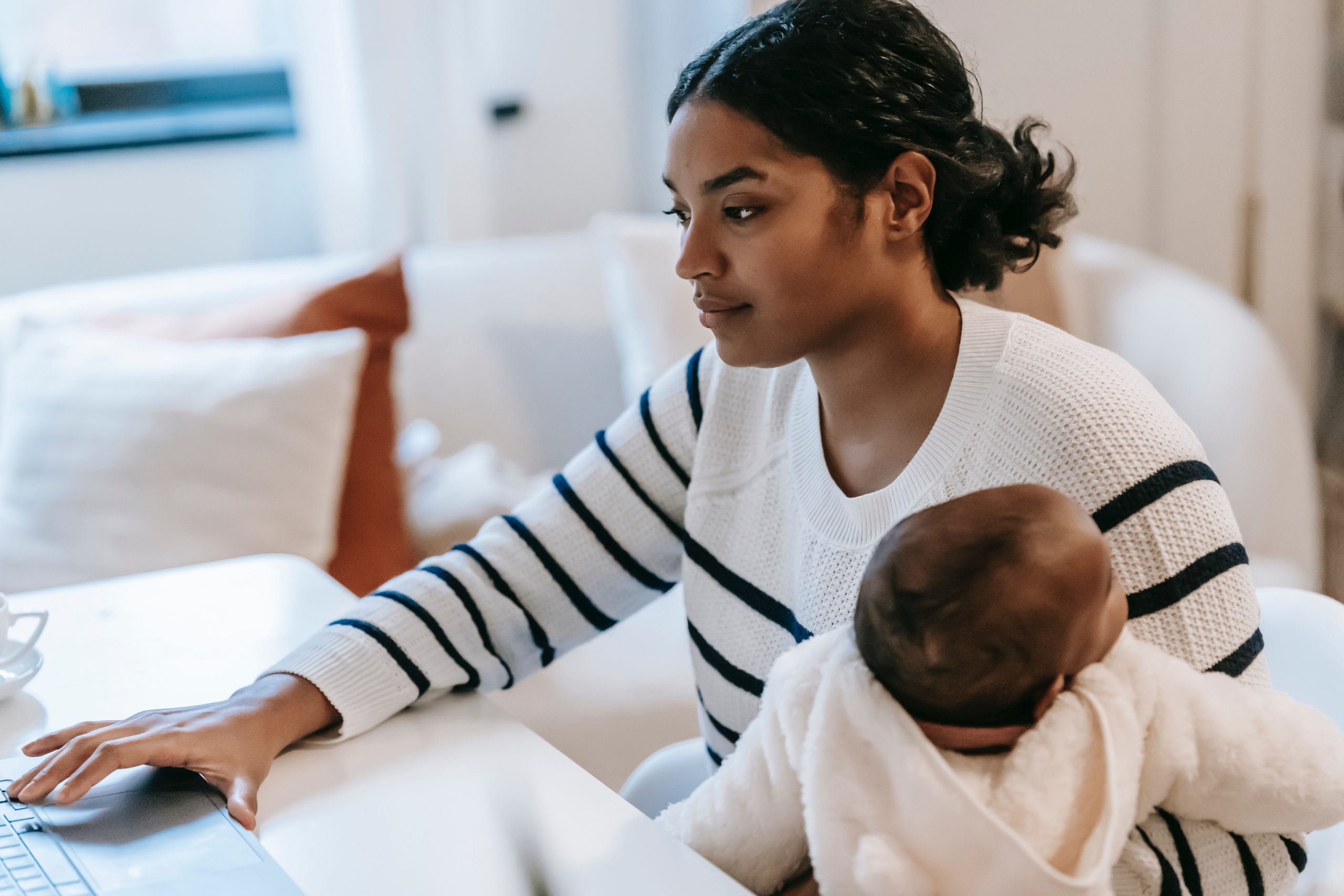 A young woman works on her laptop while looking after her baby. 