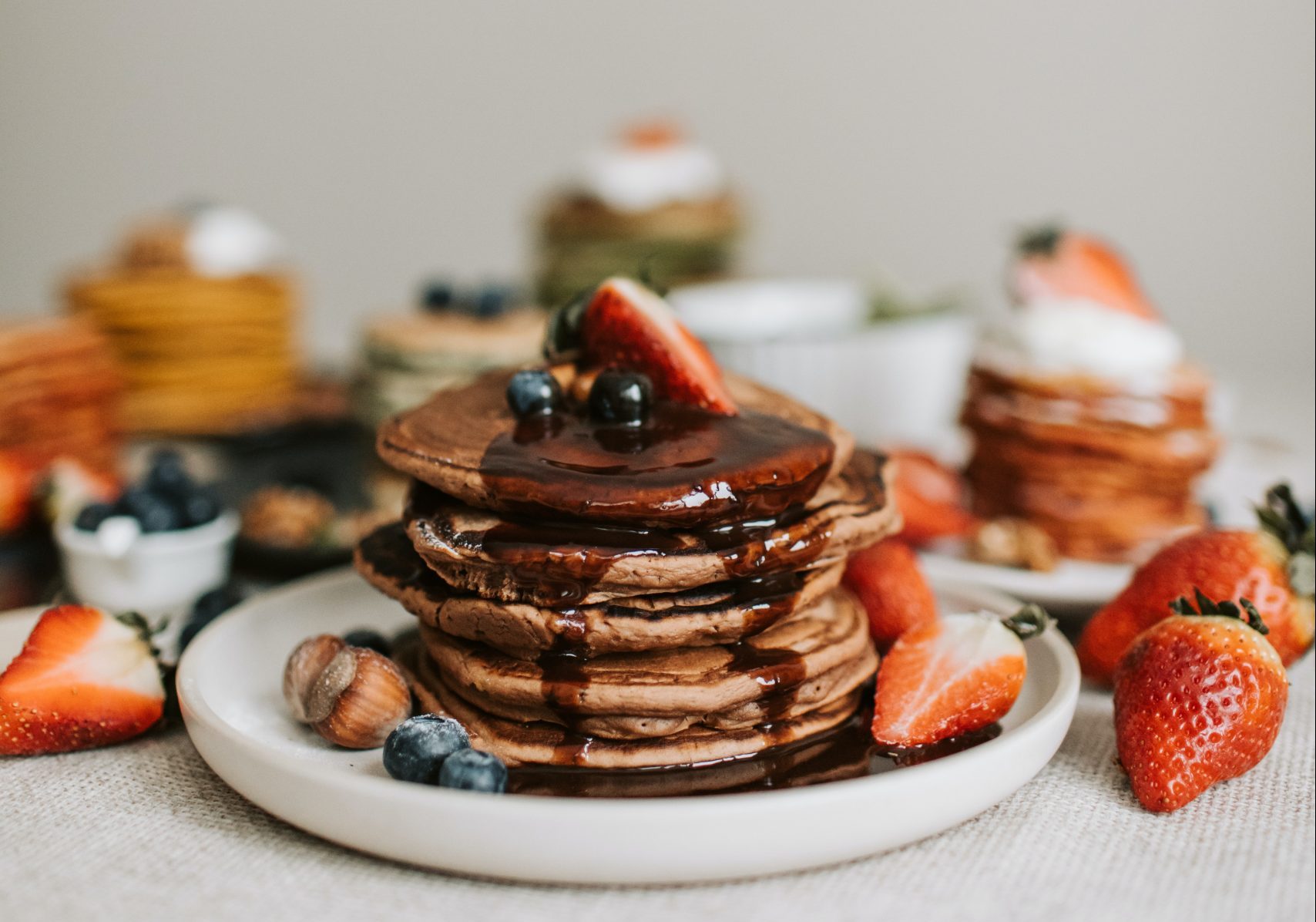 Stacks of pancakes served in several plates with brown sugar butter syrup, strawberries, blueberries and hazlenuts.