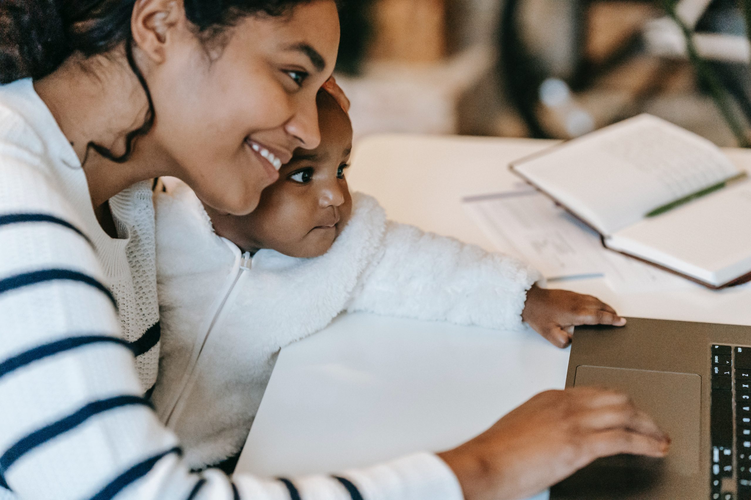 A young black woman holds her child as she works on her MacBook.