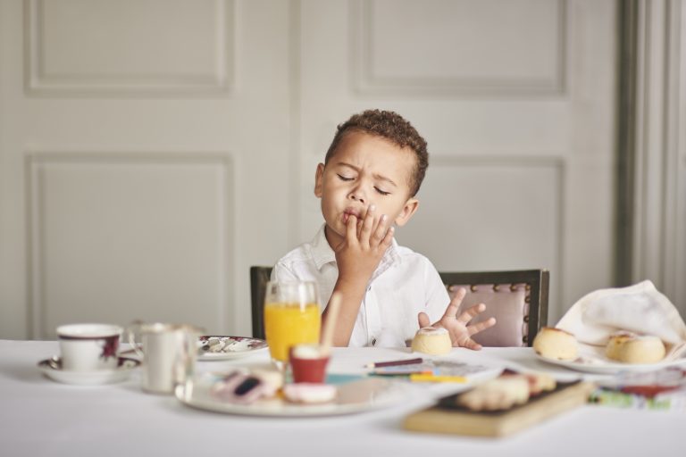 A young boy licking his fingers after tasting one of the tea cakes before him.