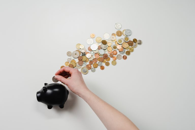 A hand placing a coin into a black piggy bank with coins streaming out from the top.