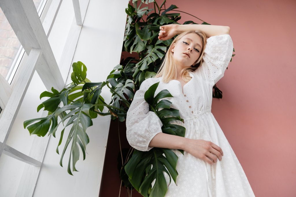 A young blonde models a white summer dress against a large green plant.