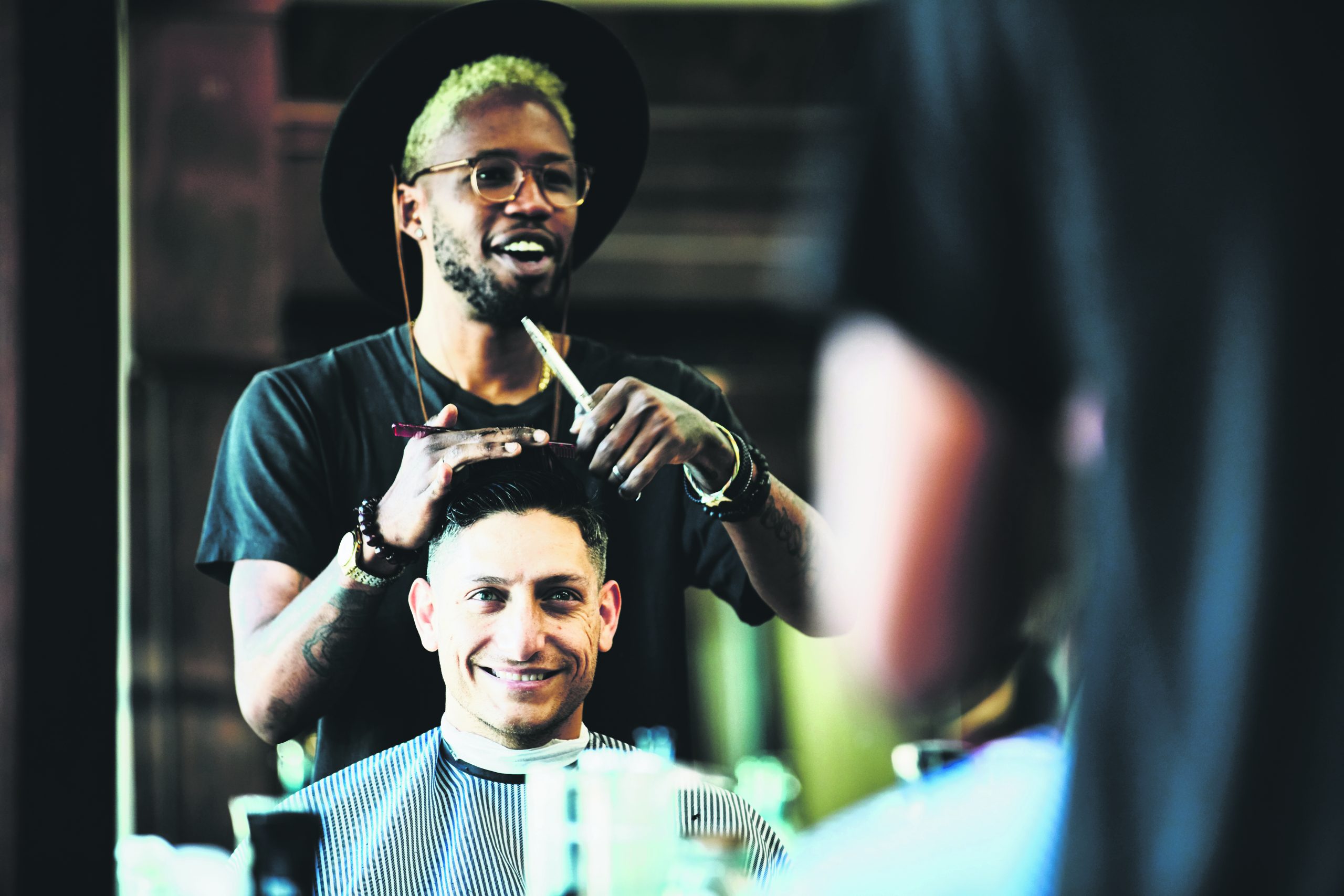 A hip, young barber bantering and joking with the young man whose hair he's cutting.