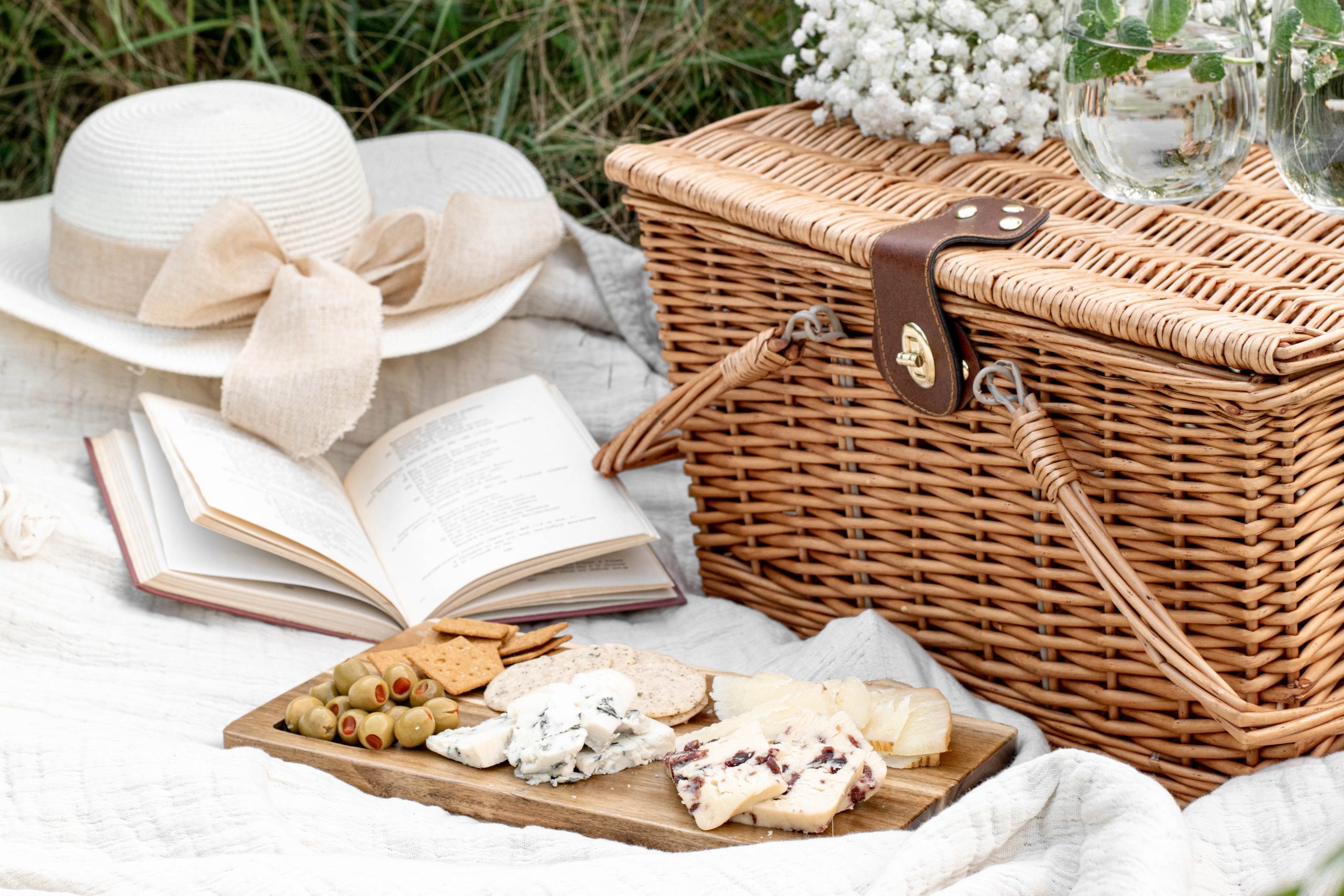 Laid out on a blanket, a cheese platter, a book, a ladies straw hat and a picnic basket.