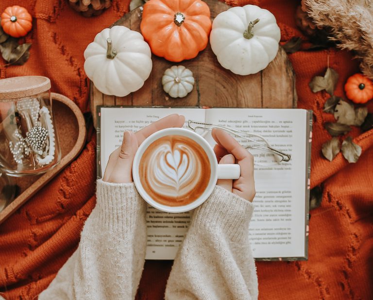A woman warming her hands on a pumpkin spiced latte while reading a book.
