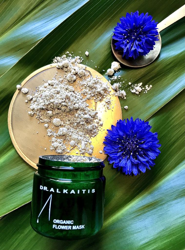 Dr Alkaitis Organic Flower Mask, an organic facial mask, resting on green Banana leaves next to blue flowers.