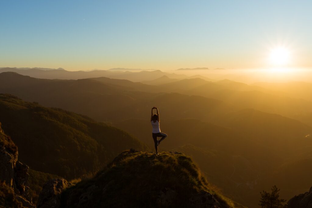 A woman practising yoga on the top of a hill as the sun sets on the horizon.