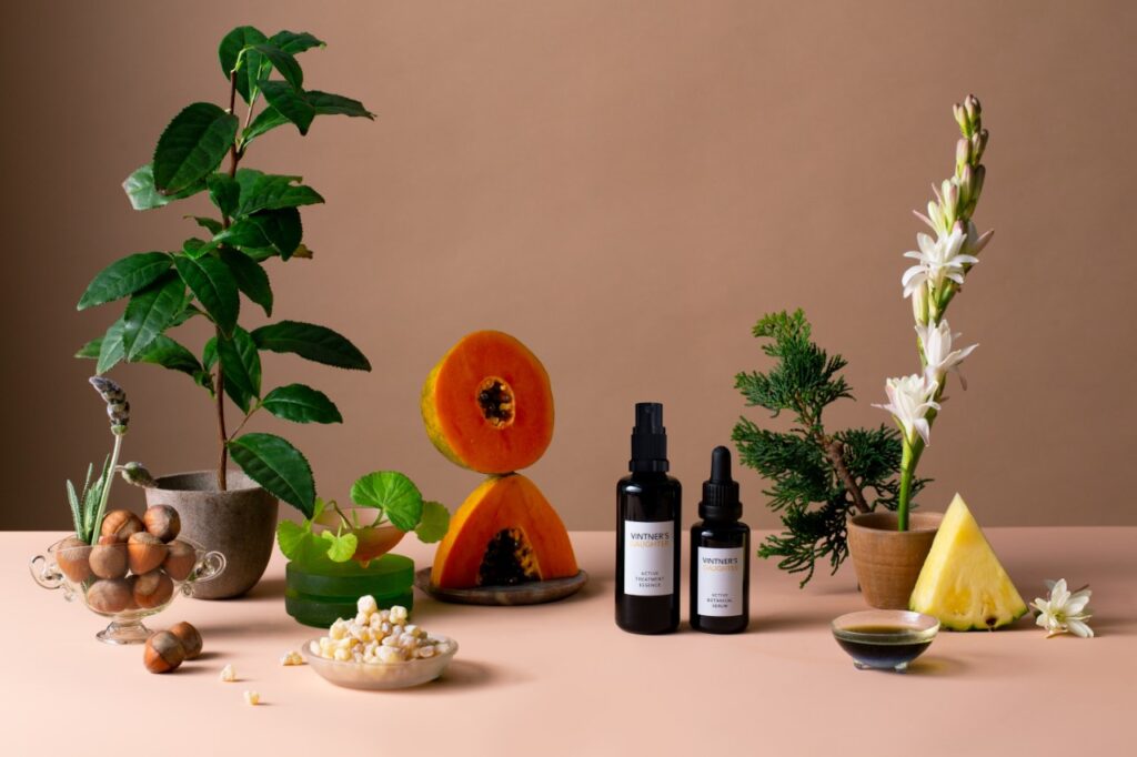 A display of two vials of Vintner’s Daughter's beauty products next to a selection of sliced fruit, green plants and bowls of nuts.
