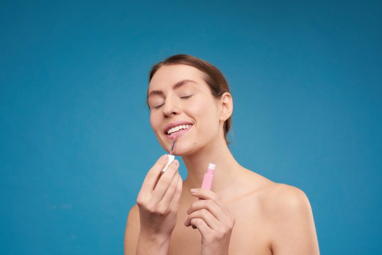 A woman applies makeup on her face against a plain blue background.