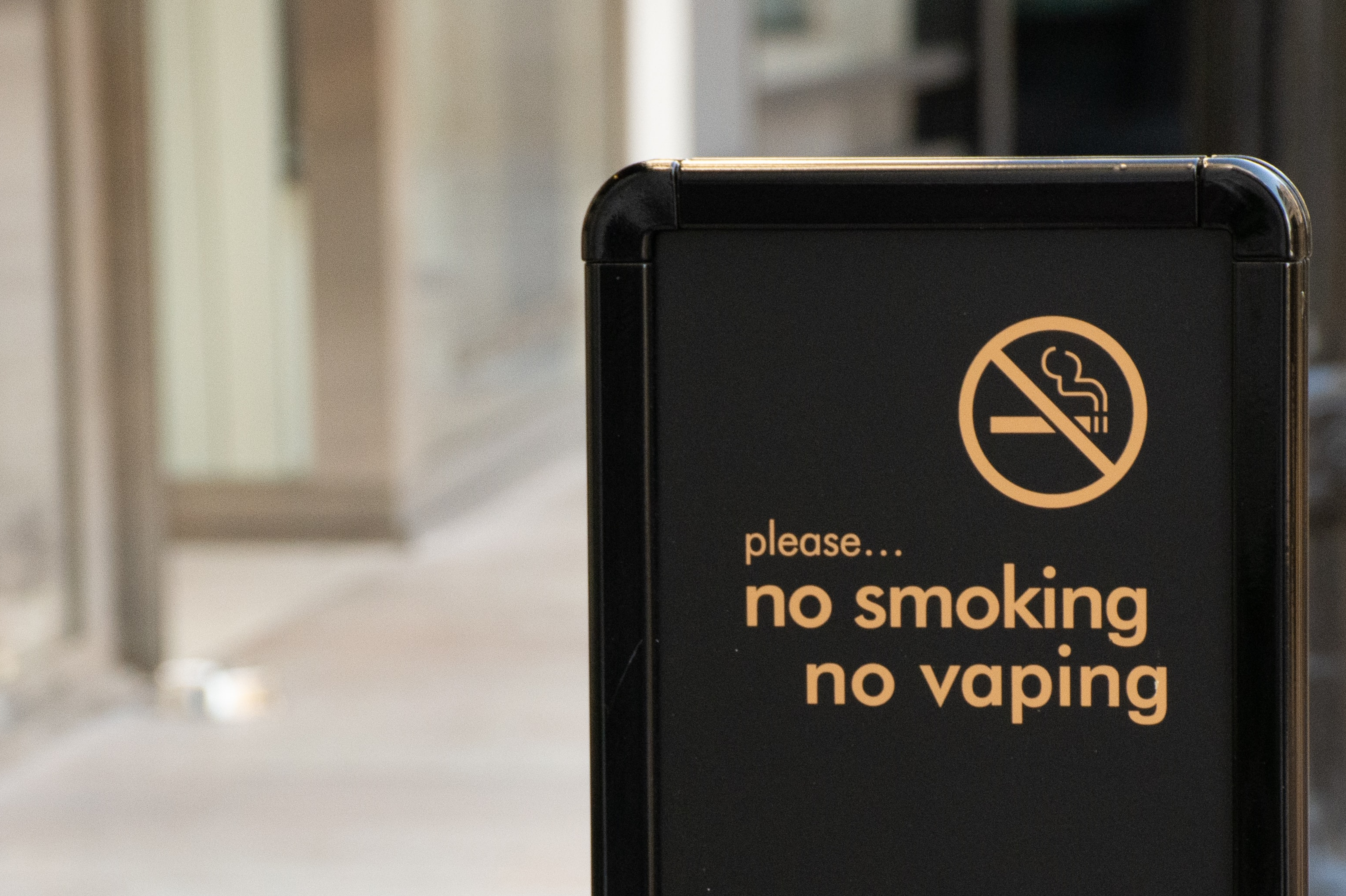 A black street sign with an anti-smoking/vaping sign and yellow letters which read, 'no smoking, no vaping'.