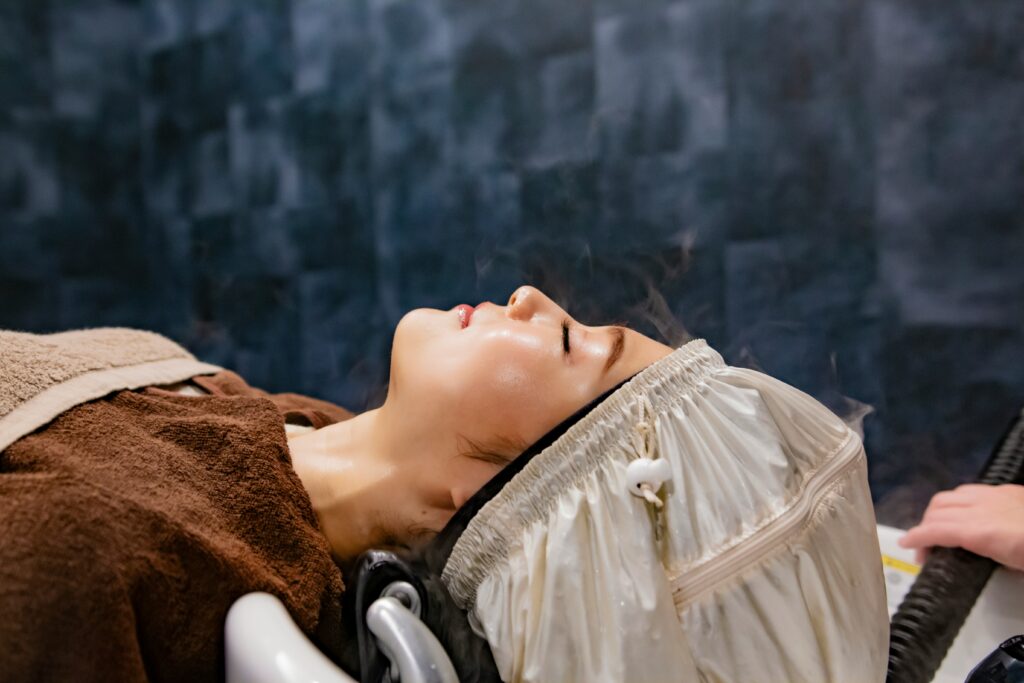 A woman lies back in a salon hair with her head being steamed