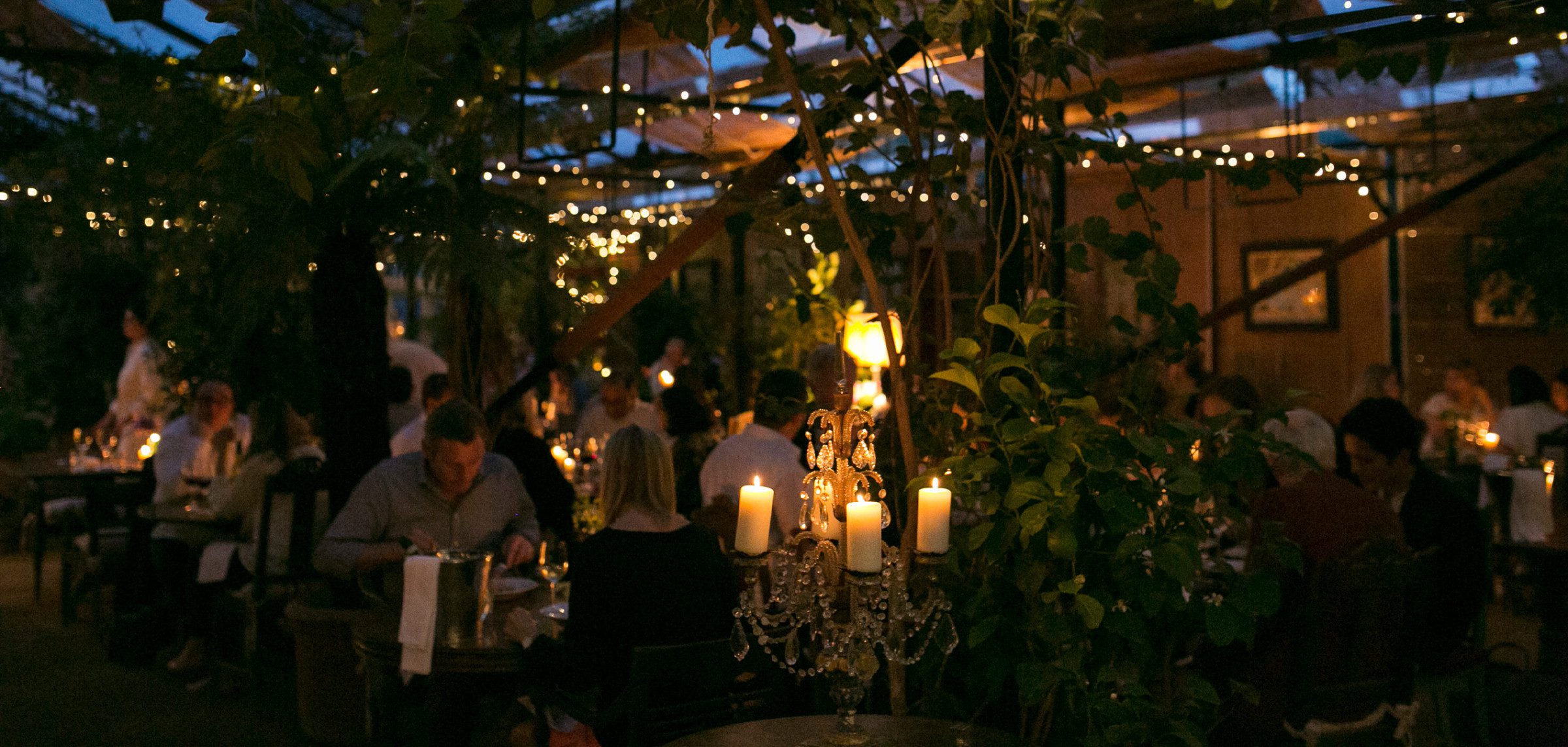 Diners enjoying food in a candle-lit greenhouse.