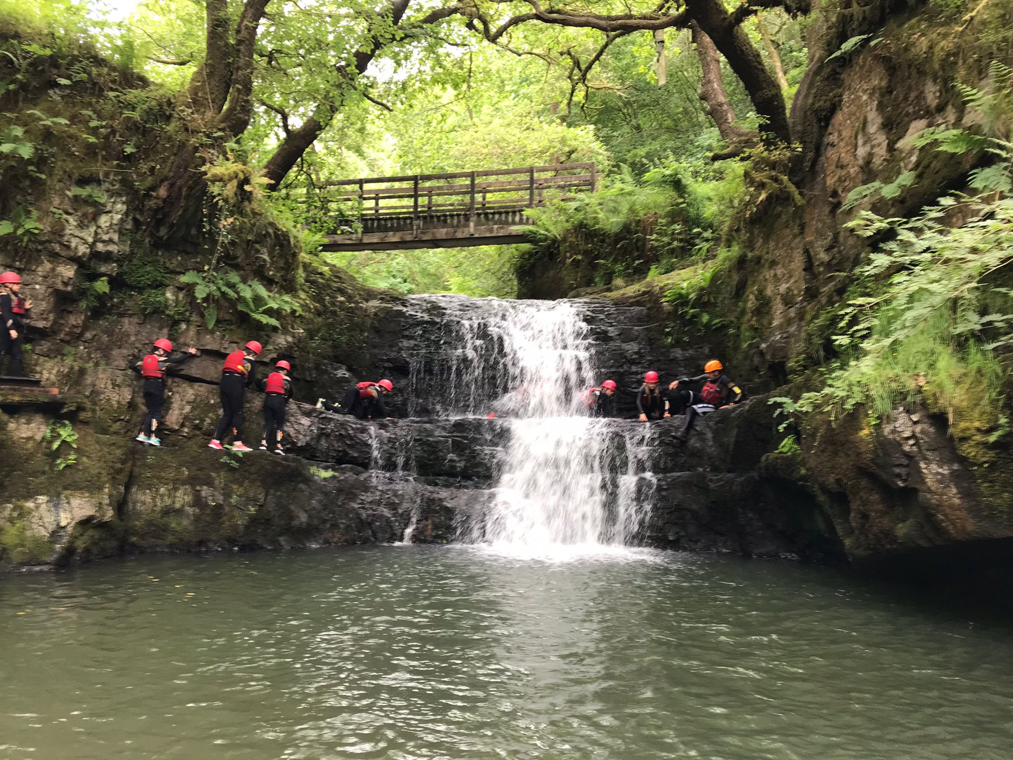 A gorge with flowing waterfall, people in helmets and life jackets walking across