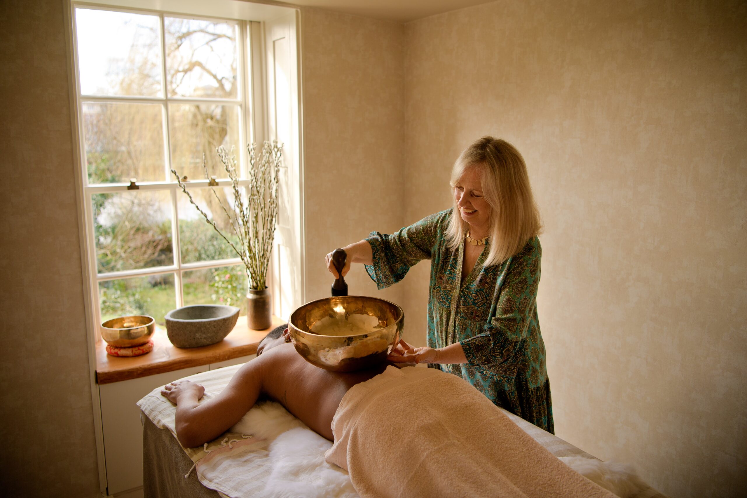 beige treatment room with window letting light in. sound healing bowls on side and man lying down on treatment bed with woman standing over ringing sound bowl gongs