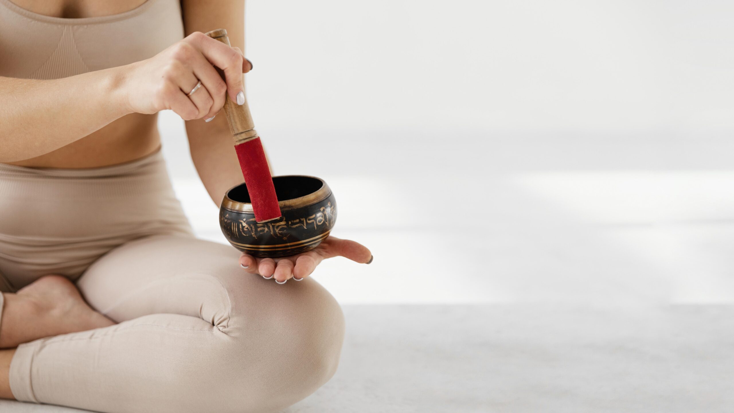 cream grey background with woman sitting cross legged in tan yoga gear holding sound healing gong