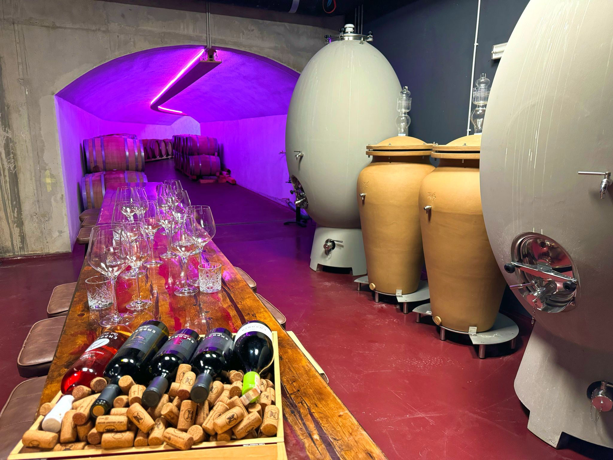 small barrel room with egg shaped barrels a long table with wine glasses and a box of wine bottles and corks