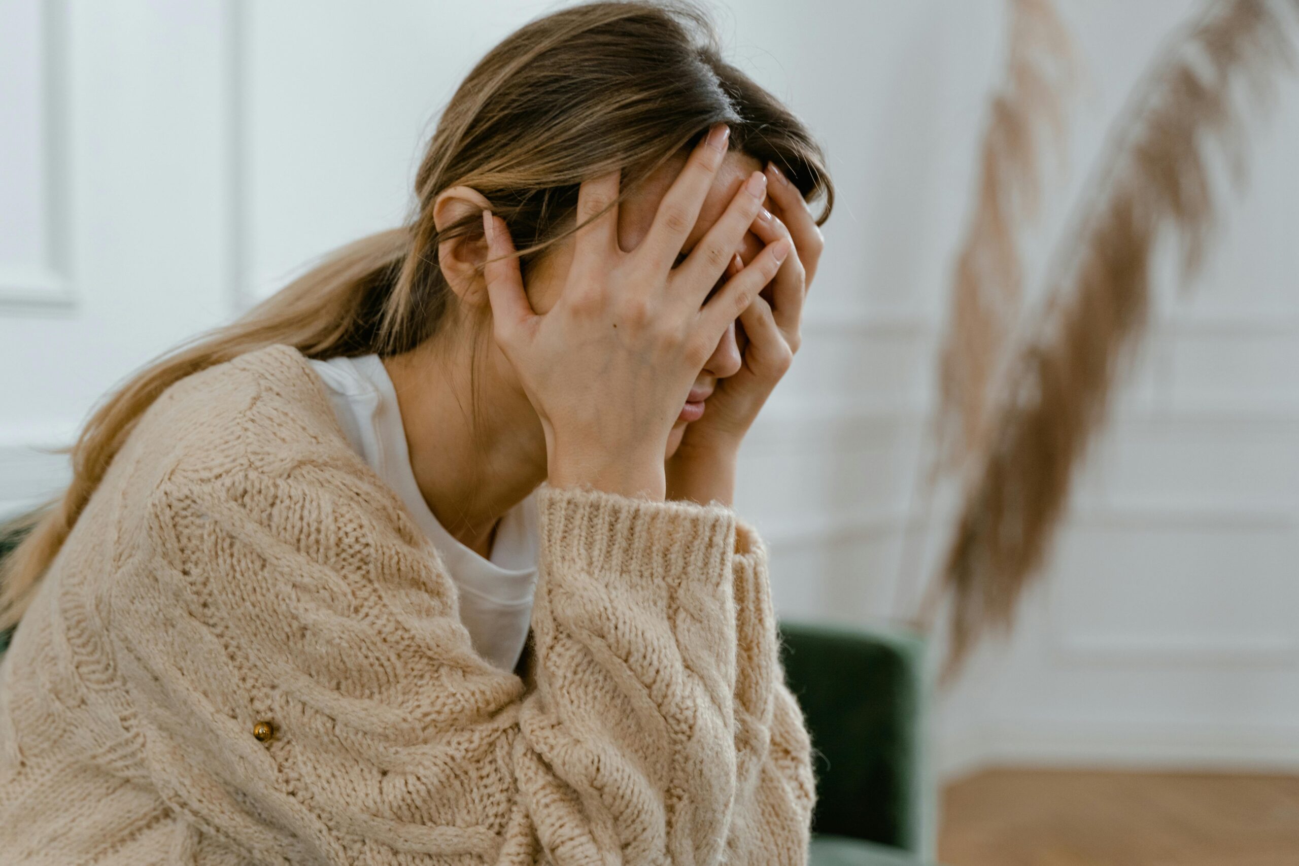 woman sits on sofa with beige jumper with her hands over her face